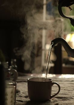 Steam rises as hot tea is elegantly poured into a mug from a kettle.