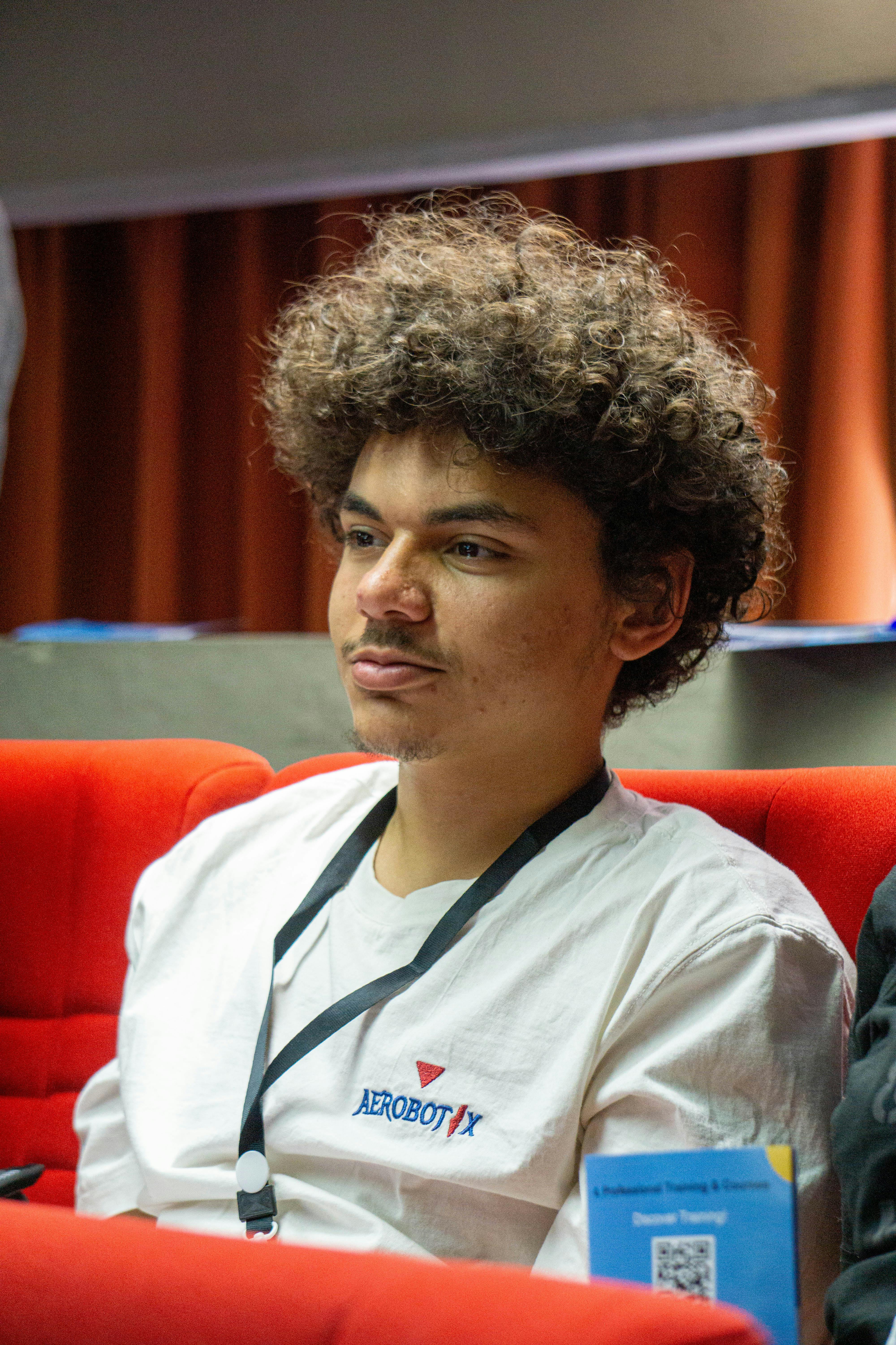 Young man with curly hair in white t-shirt attending a conference, sitting on a red chair indoors.