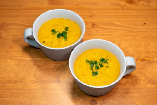 Delicious homemade pumpkin soup served in mugs, garnished with fresh parsley, on a wooden table.