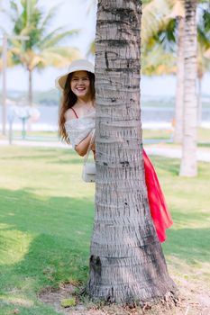 Smiling woman in a sun hat peeking from behind a palm tree in a sunny park setting.
