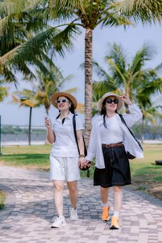 Two women walk hand in hand in a sunny park, wearing hats and sunglasses.