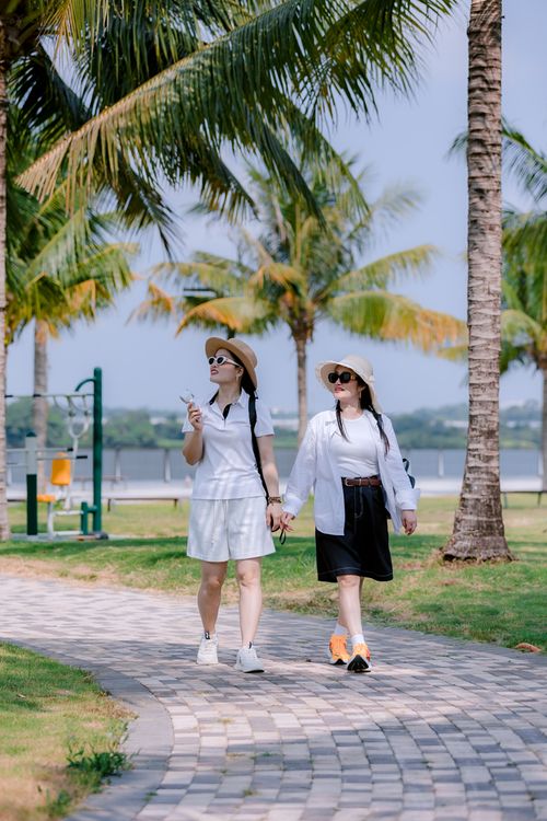 Free Two women walking hand in hand through a palm tree-lined park path on a sunny day. Stock Photo