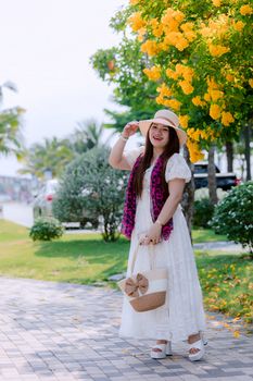 A smiling woman in a white dress and sun hat enjoys a sunny walk under vibrant yellow blooms.