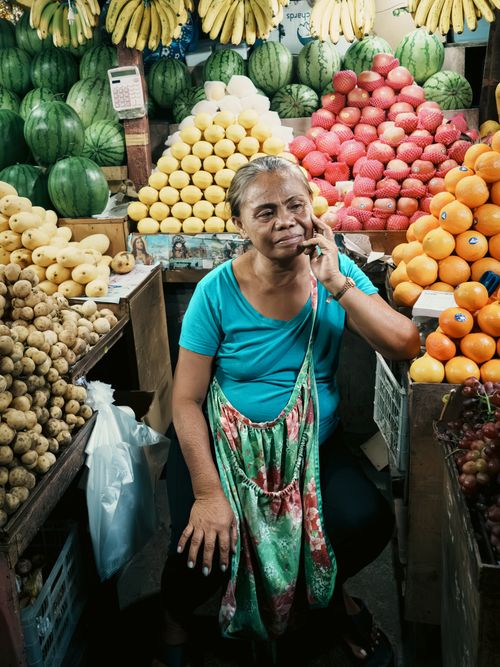 Free A female vendor at a colorful fruit stand in Cebu City, Philippines, engaged in a phone call surrounded by fresh produce. Stock Photo