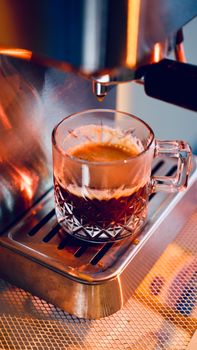 Close-up of espresso in crystal glass cup emerging from coffee machine.