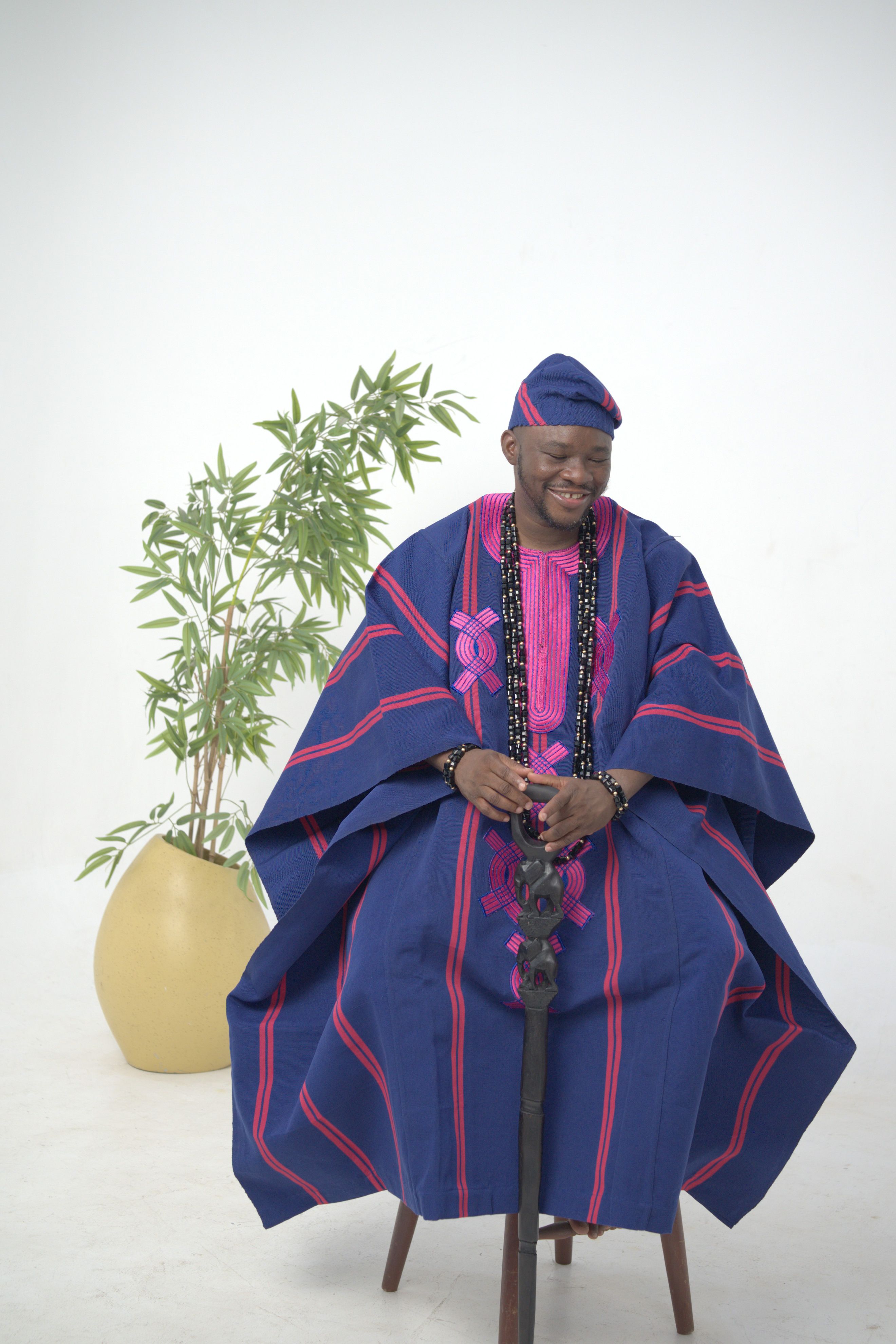 Man in traditional blue and pink Nigerian attire seated indoors with a plant.