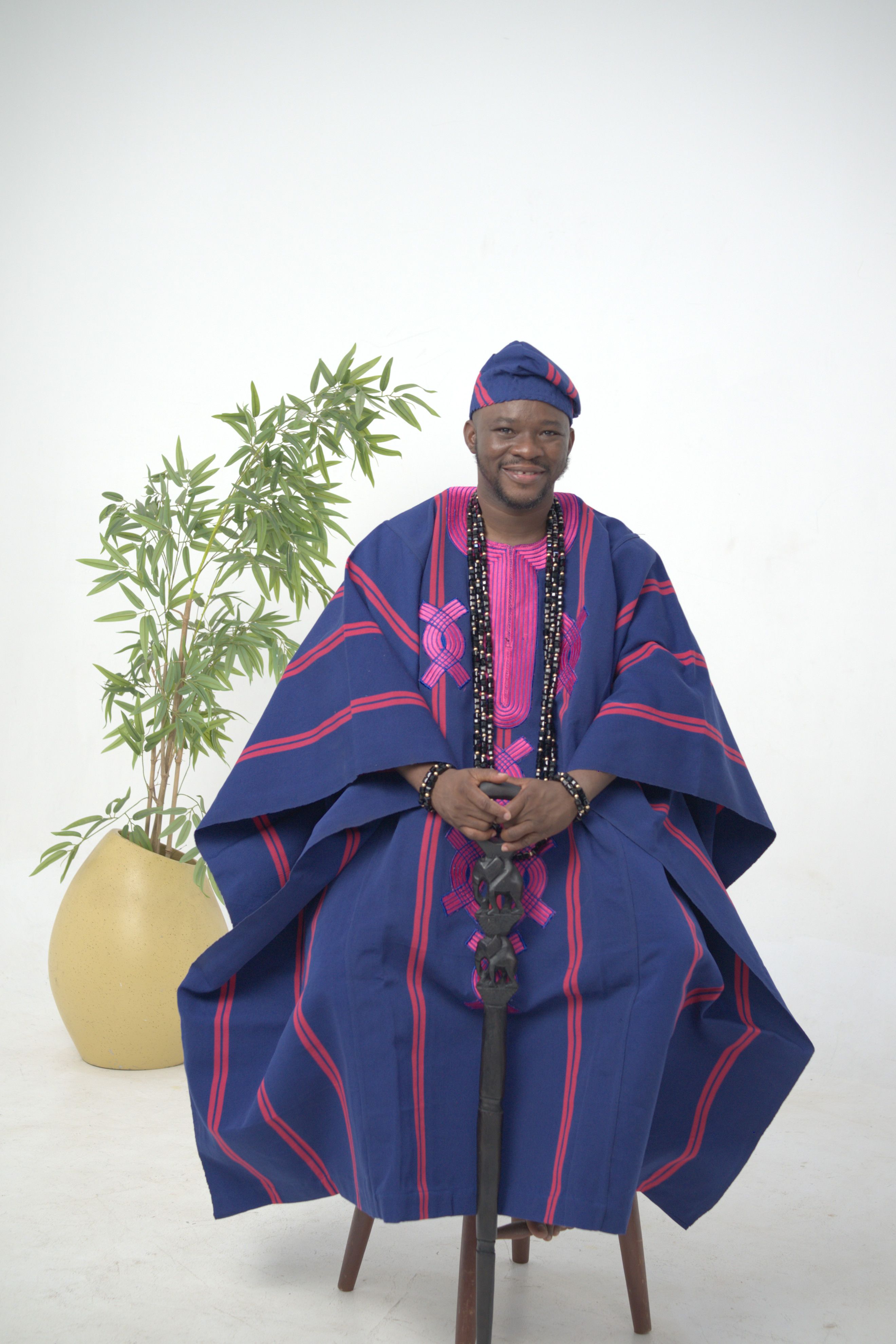Portrait of a man in traditional Nigerian attire, seated indoors with a plant.