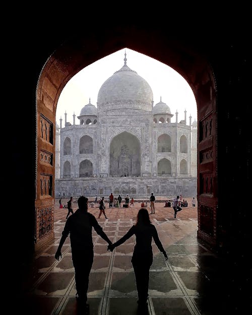 Back view of silhouette of loving traveling couple holding hands and walking through arch towards Taj Mahal