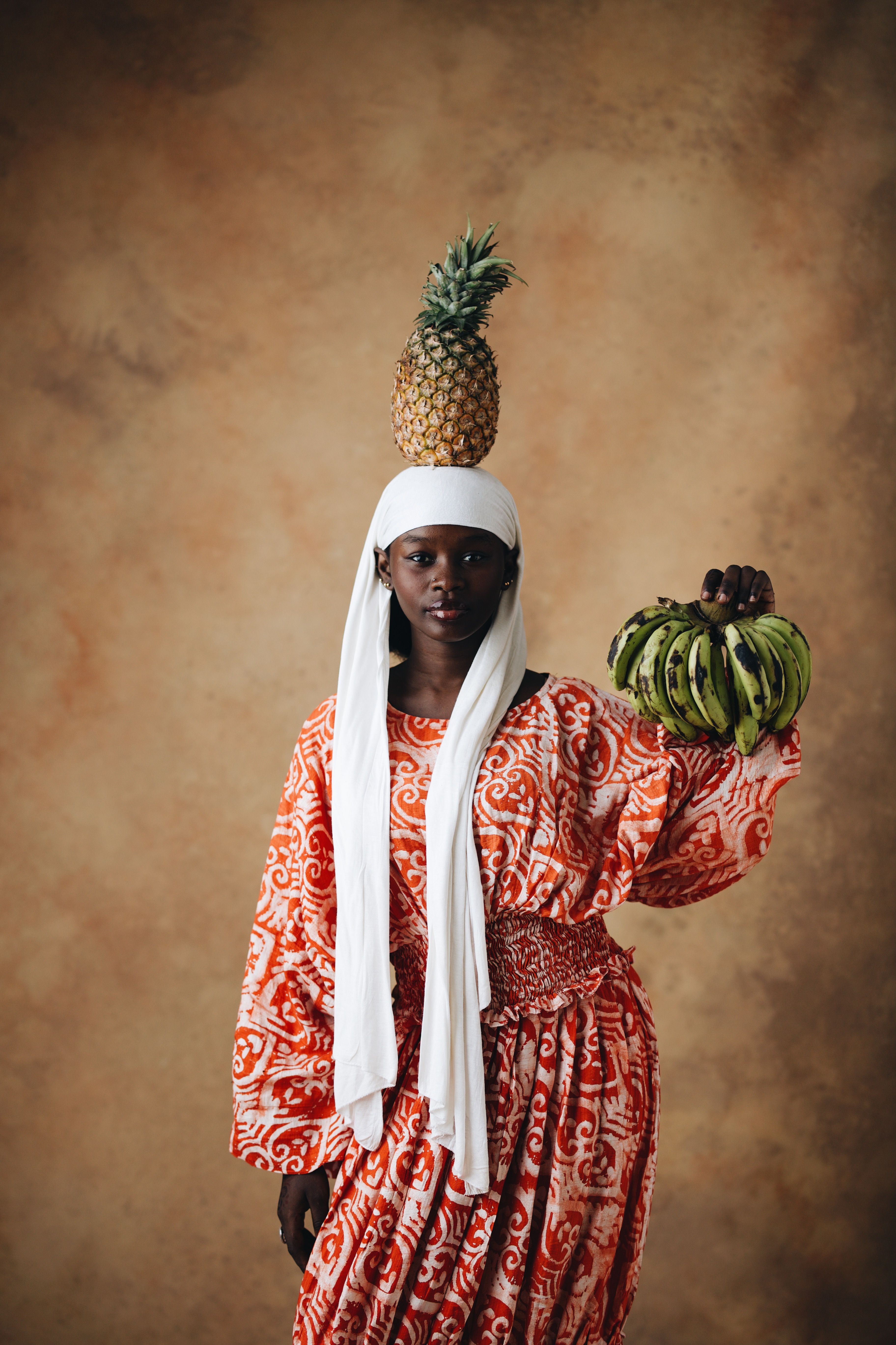 Nigerian woman in cultural attire balancing pineapple with bananas, showcasing traditional art.