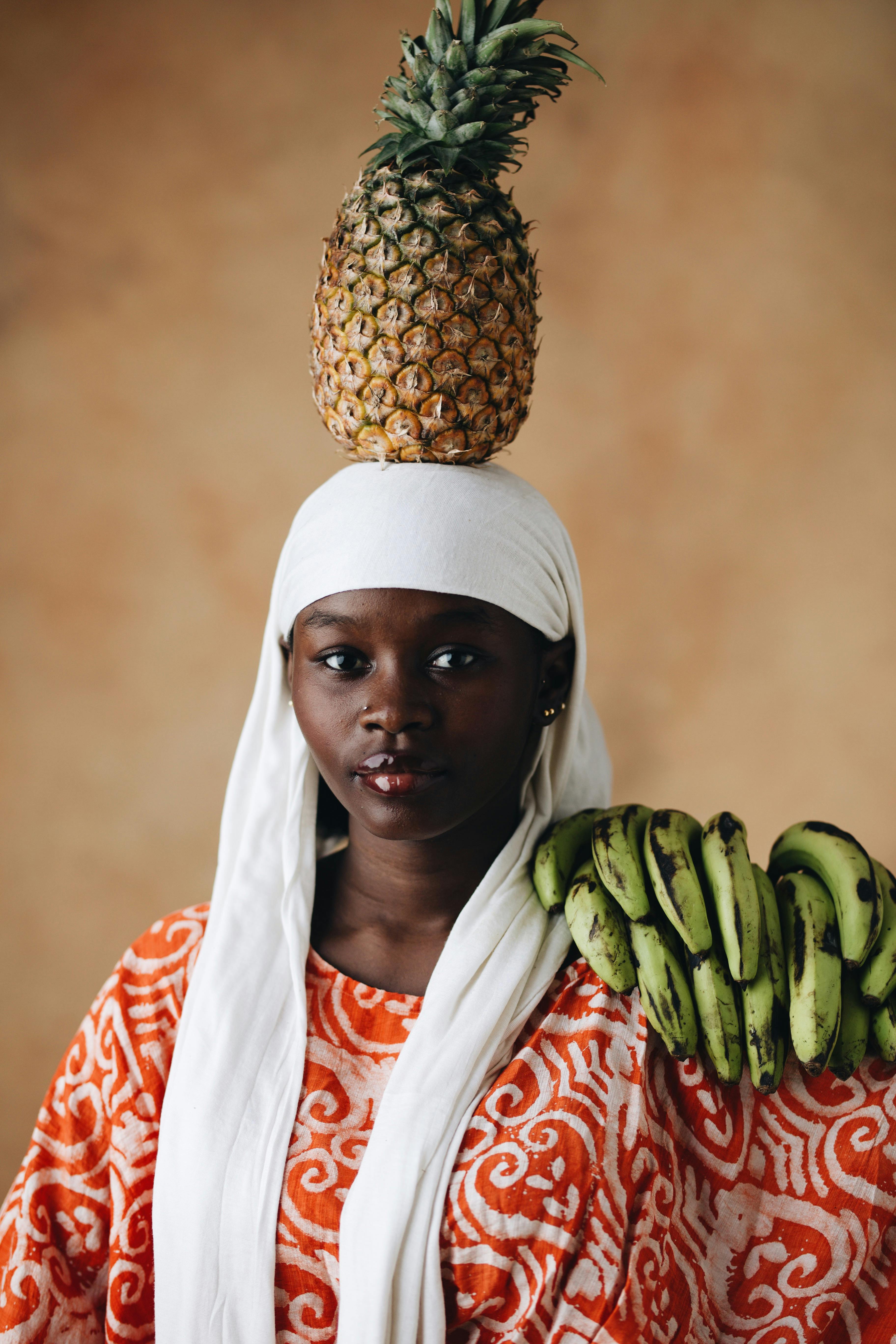 African woman balancing a pineapple on her head while holding bananas, showcasing traditional attire.