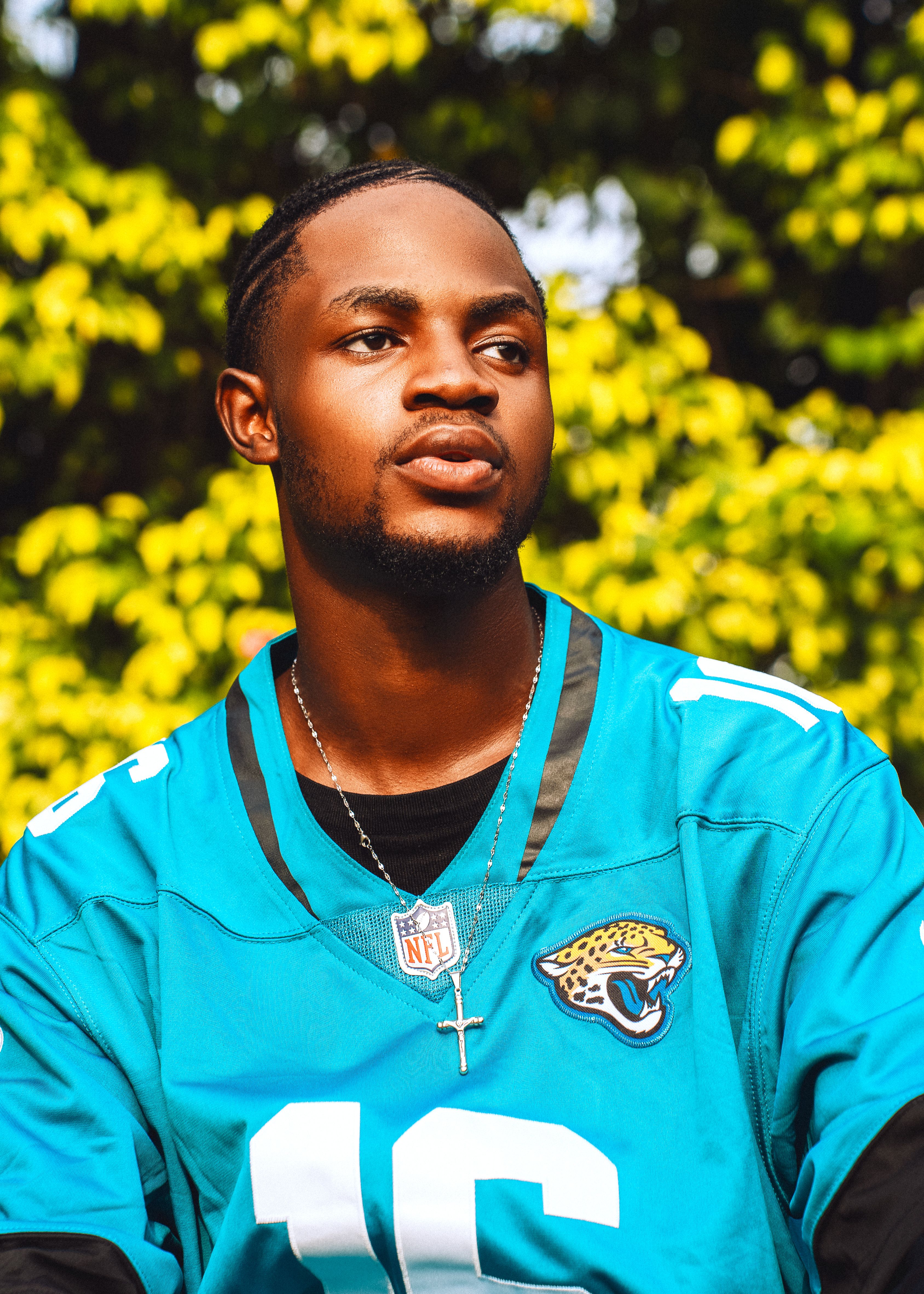 Portrait of a young man wearing a teal jersey with a cross necklace, set against a vibrant autumn backdrop.