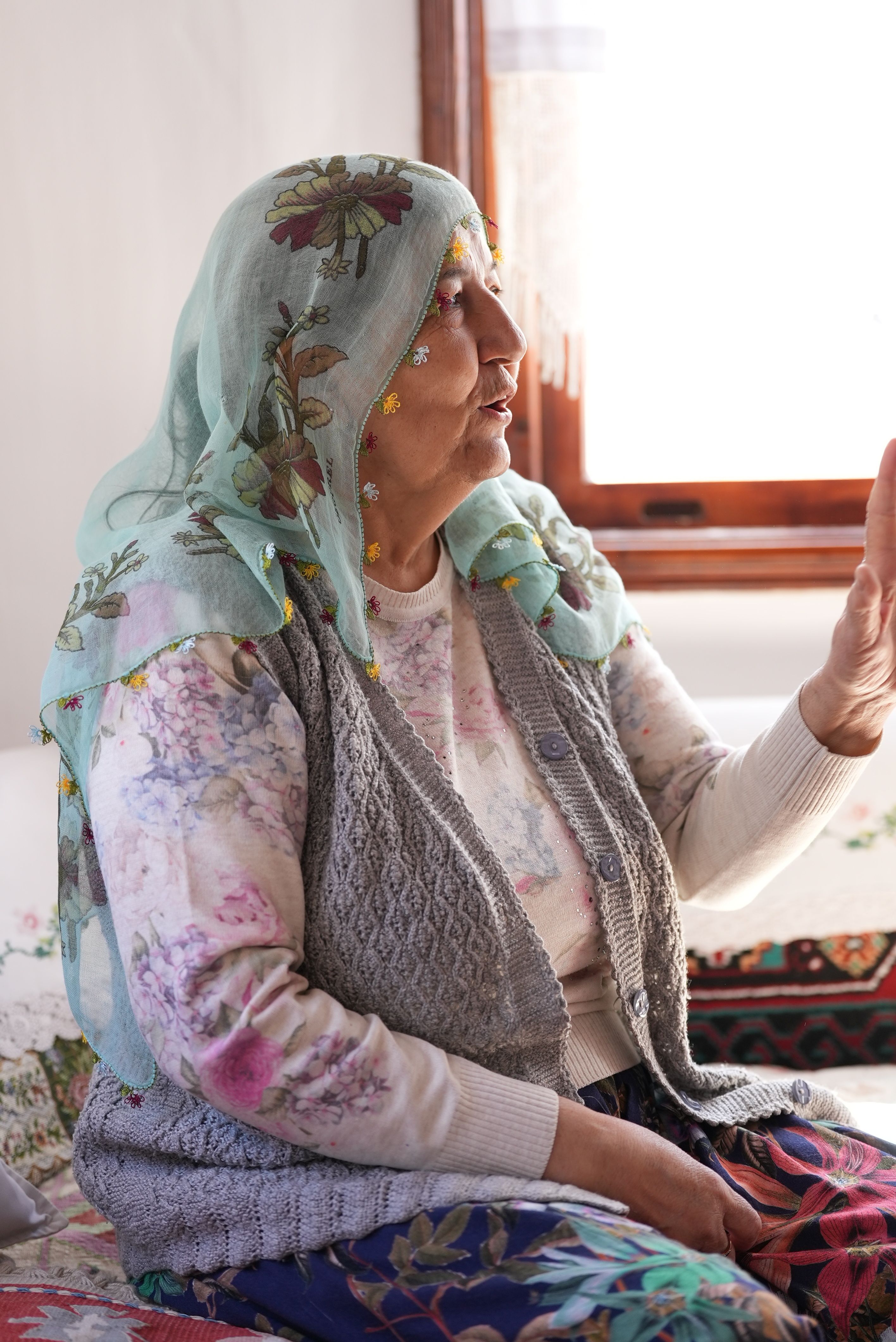 Elderly woman wearing a floral embroidered headscarf, sitting indoors near a window.