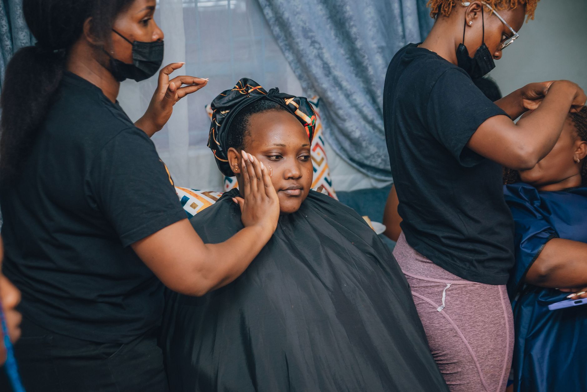 A bride receives makeup and styling assistance before her wedding in an indoor setting.