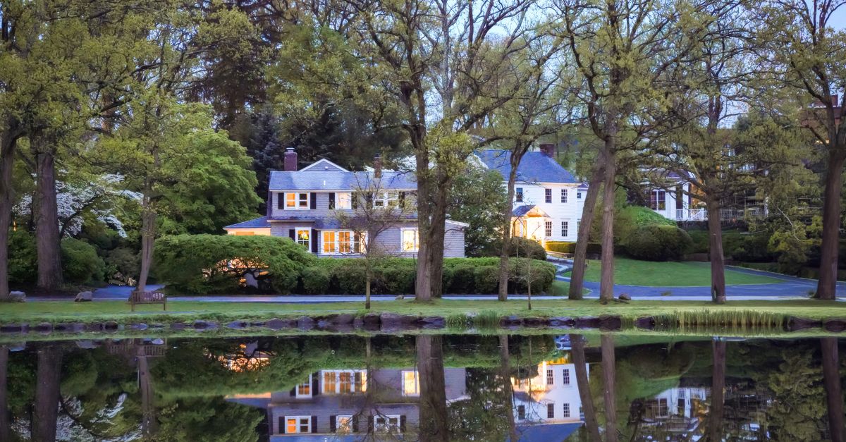 Charming suburban house reflected in a serene pond surrounded by lush trees at twilight.