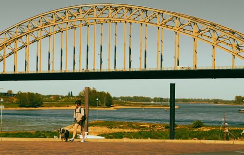 Free A woman walks her dog by a large arch bridge over a river on a sunny day. Stock Photo