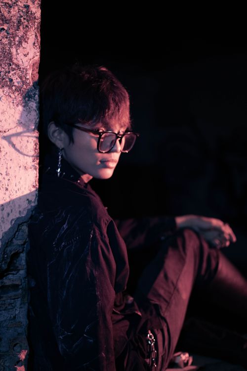 Free A stylish young adult wearing glasses sitting against a textured wall in dramatic lighting. Stock Photo