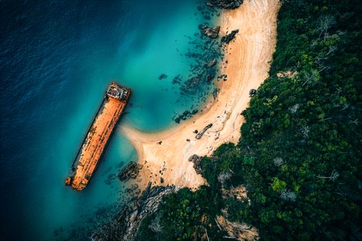Stunning aerial view of a shipwreck on a deserted tropical beach with lush greenery and clear blue water.