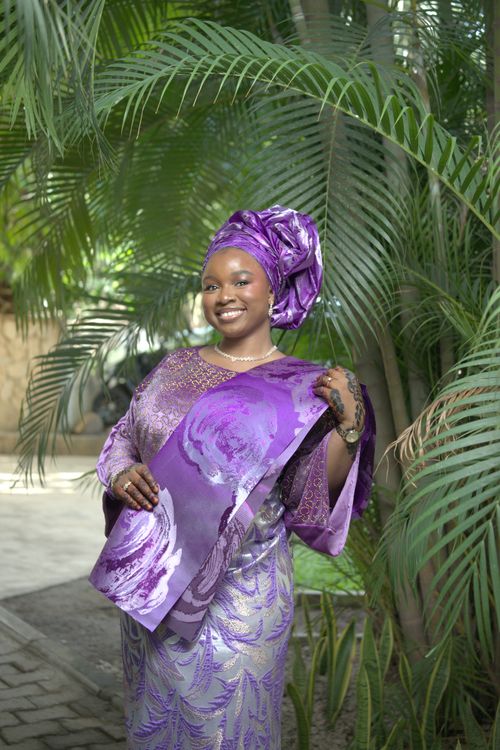 Free Smiling Nigerian woman wearing purple Ashoke attire in a lush outdoor setting. Stock Photo