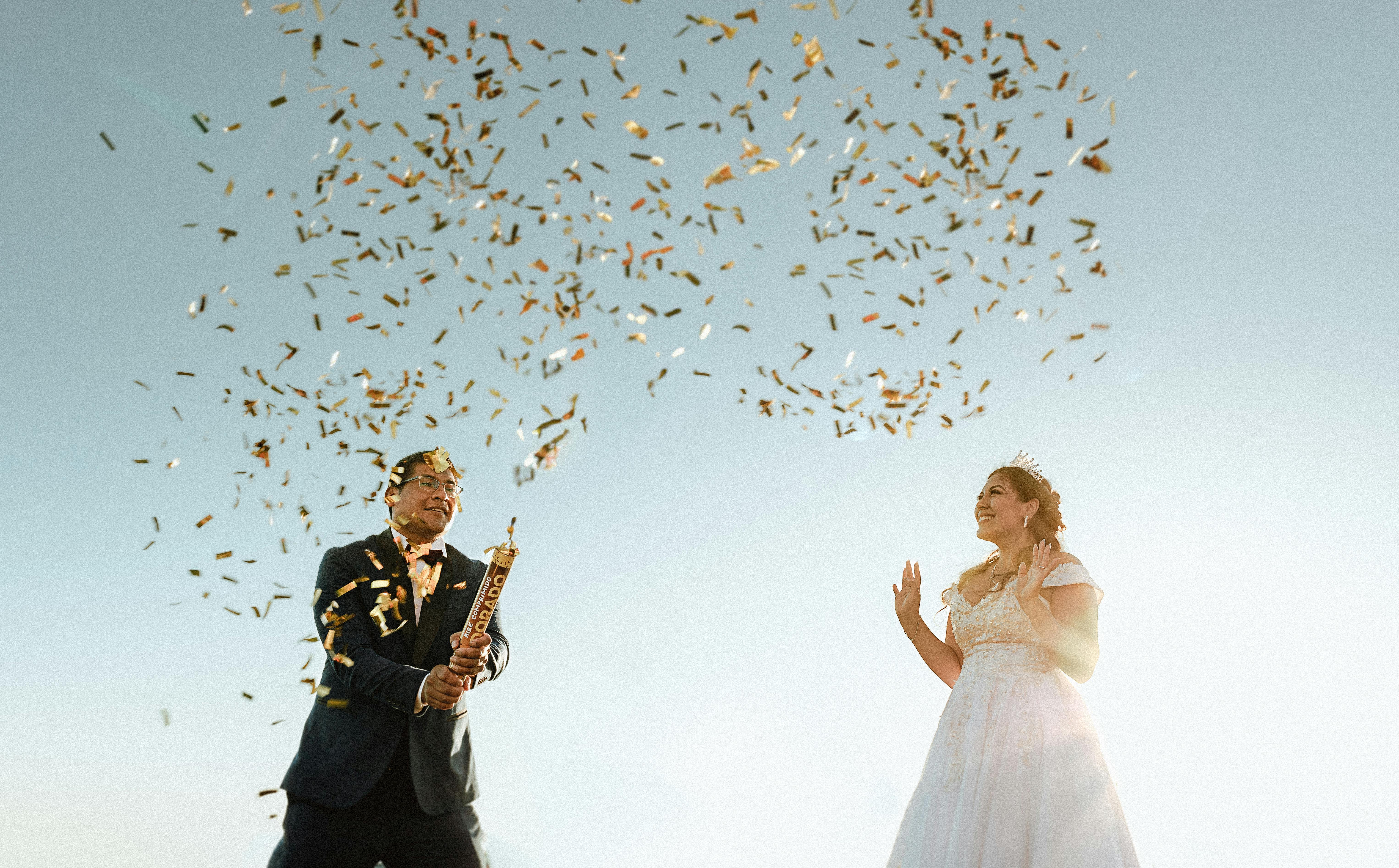 Happy bride and groom celebrate outdoors with confetti explosion under a clear sky.