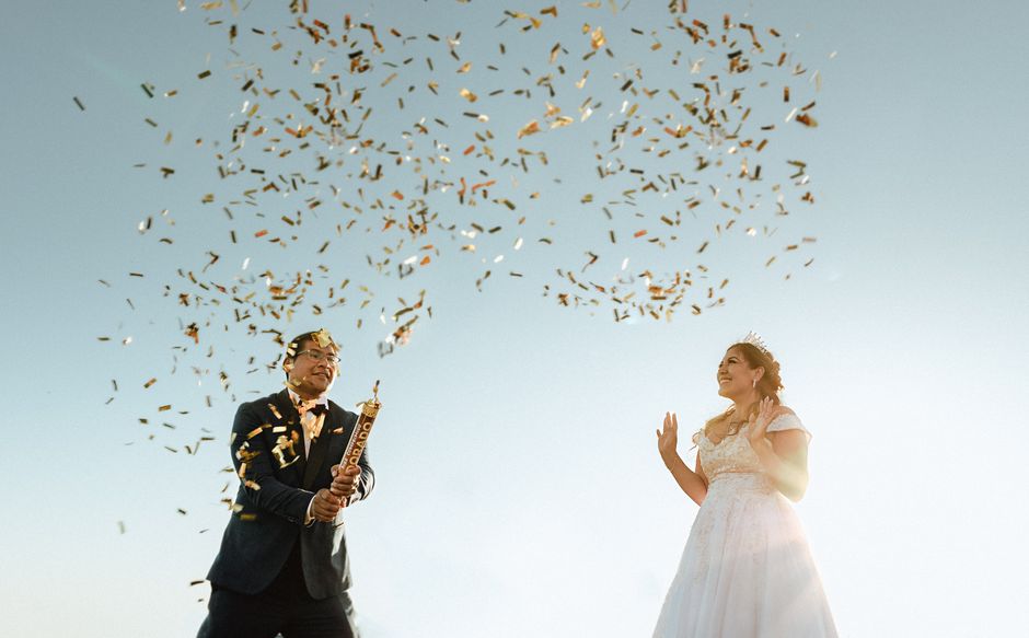 Happy bride and groom celebrate outdoors with confetti explosion under a clear sky.