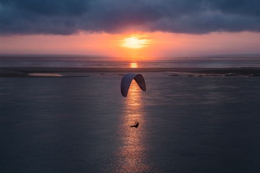 Paraglider silhouette soaring over calm waters with a stunning sunset backdrop, capturing adventure and tranquility.