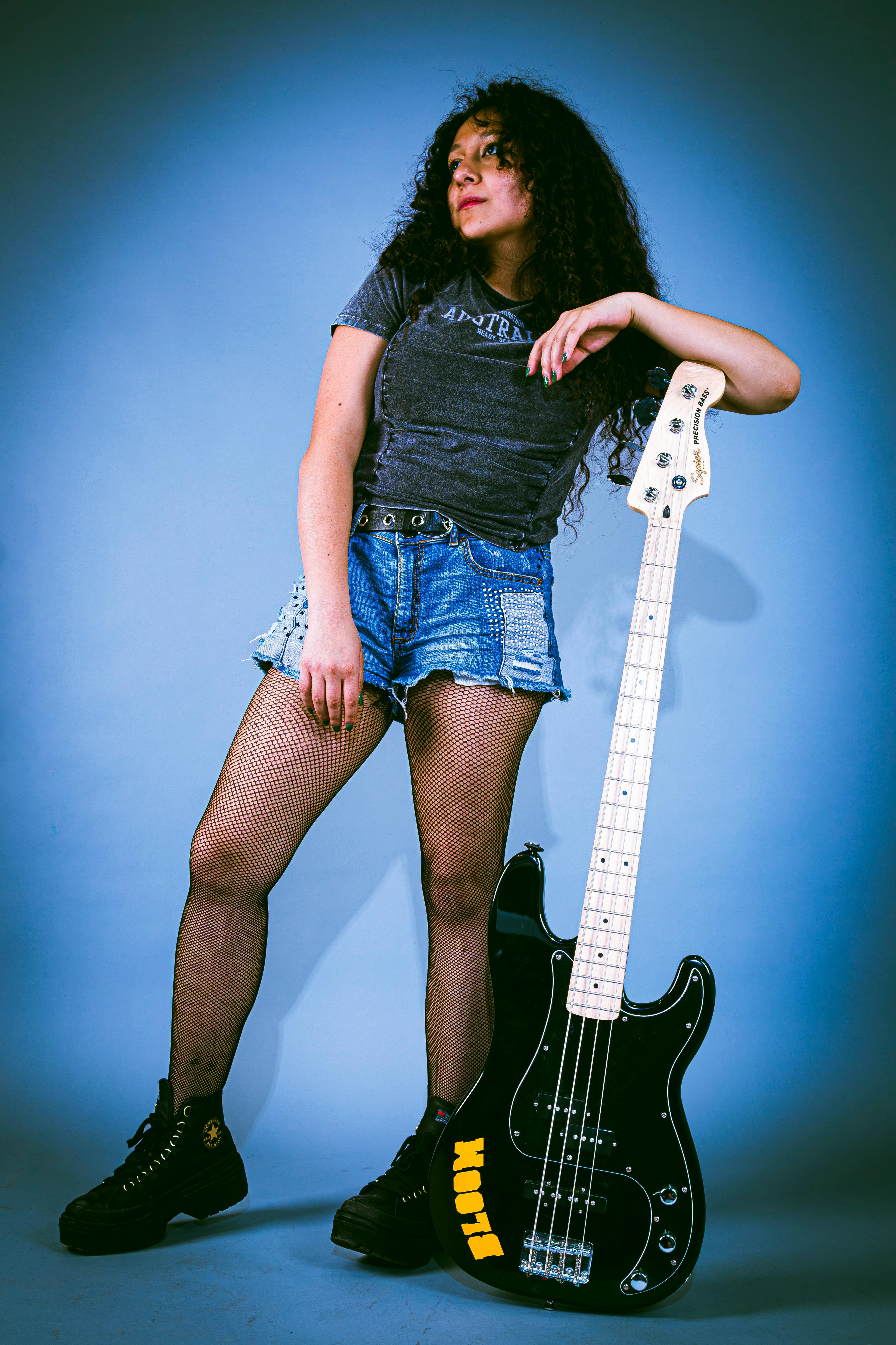 Confident young woman posing with an electric bass guitar against a blue background.