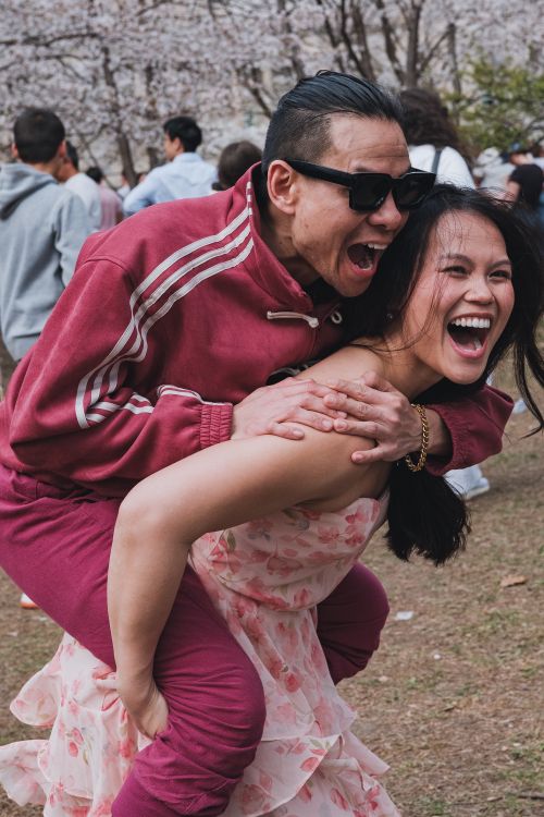 Free A happy couple shares a playful piggyback ride outdoors, surrounded by people and trees. Stock Photo
