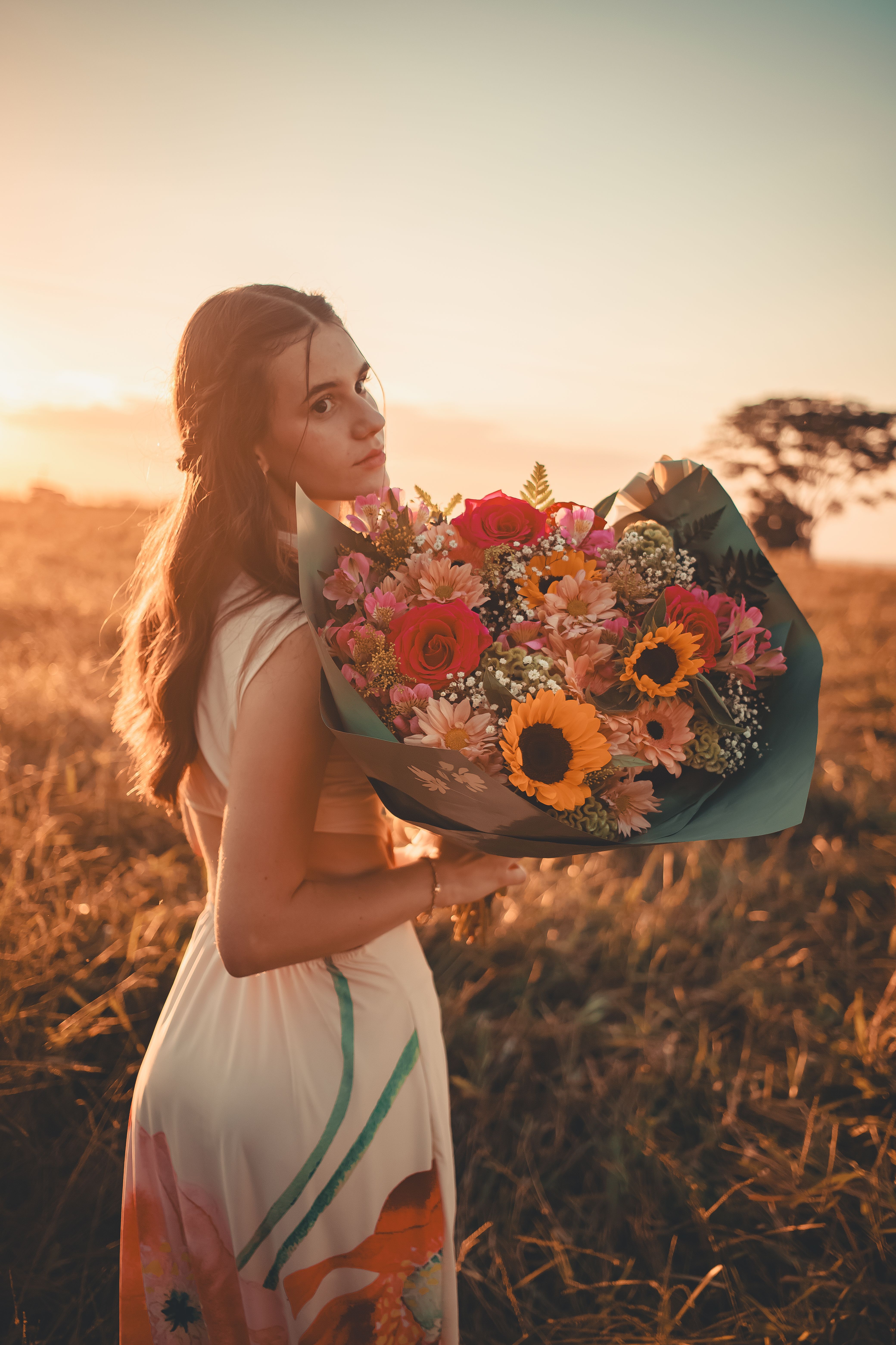A young woman stands in a field with a colorful flower bouquet during sunset, embracing nature's beauty.
