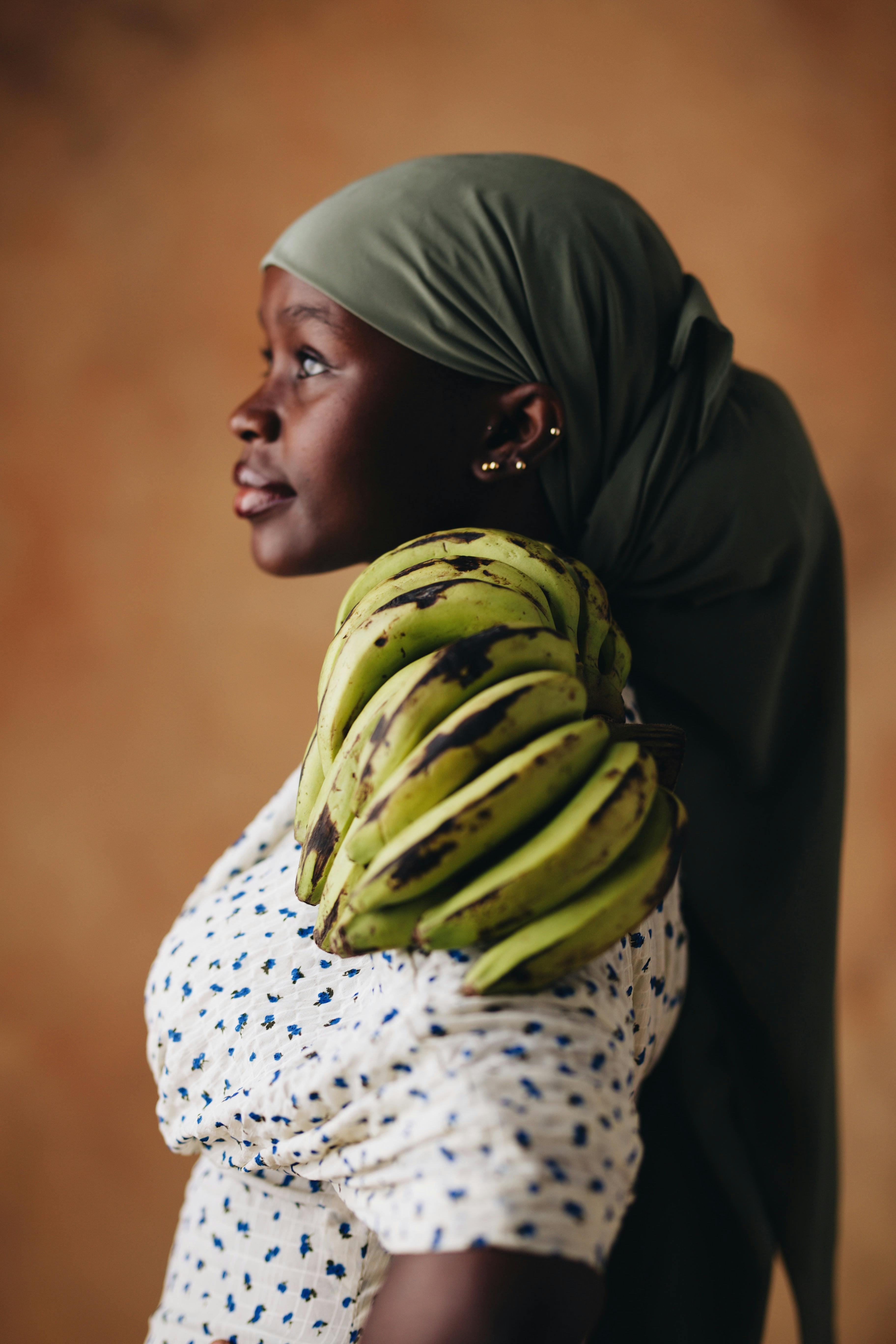 Woman in headscarf carries green plantains on shoulder, side profile.