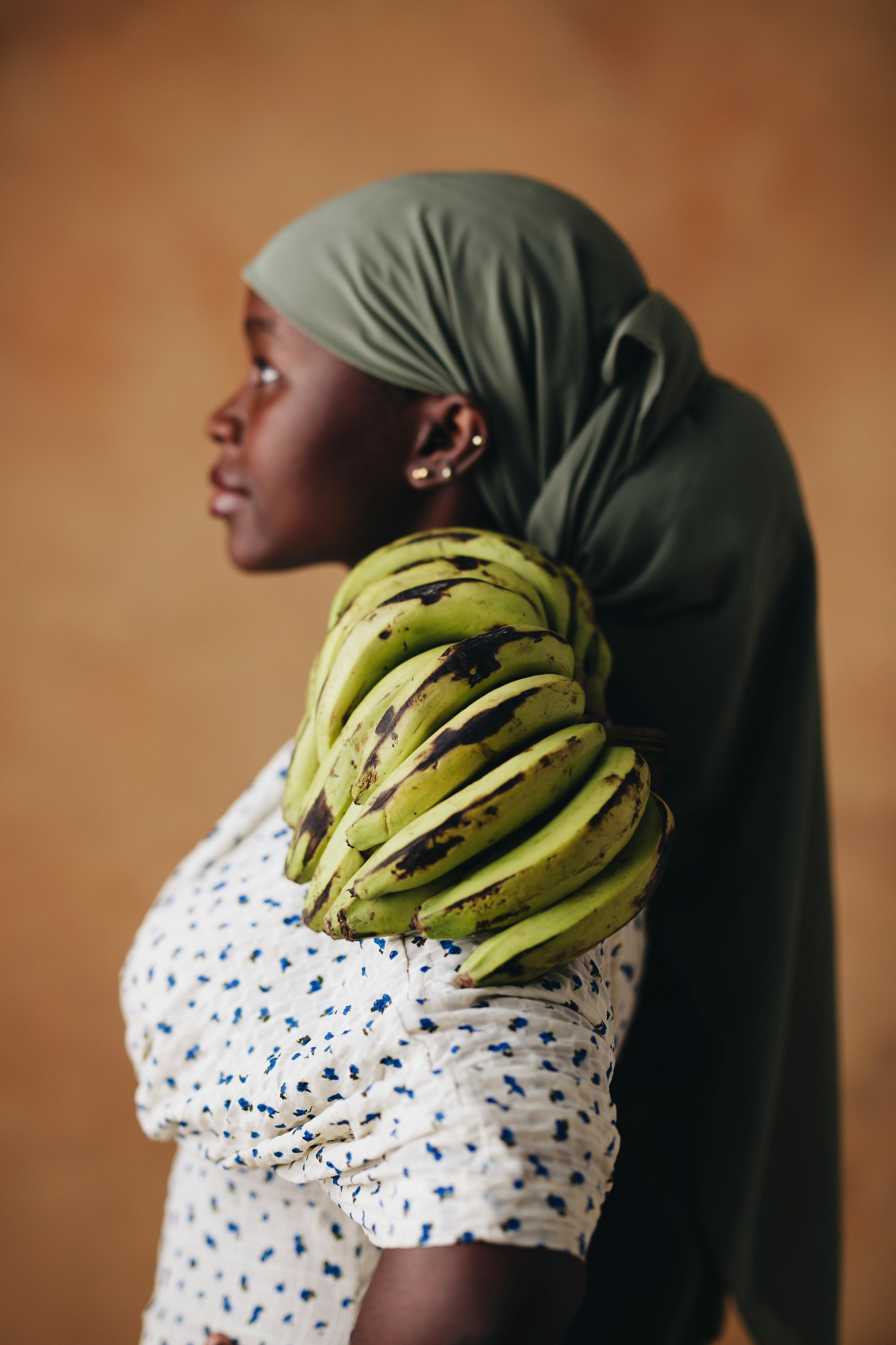 Profile portrait of an African woman wearing a headscarf, carrying green plantains on her shoulder.