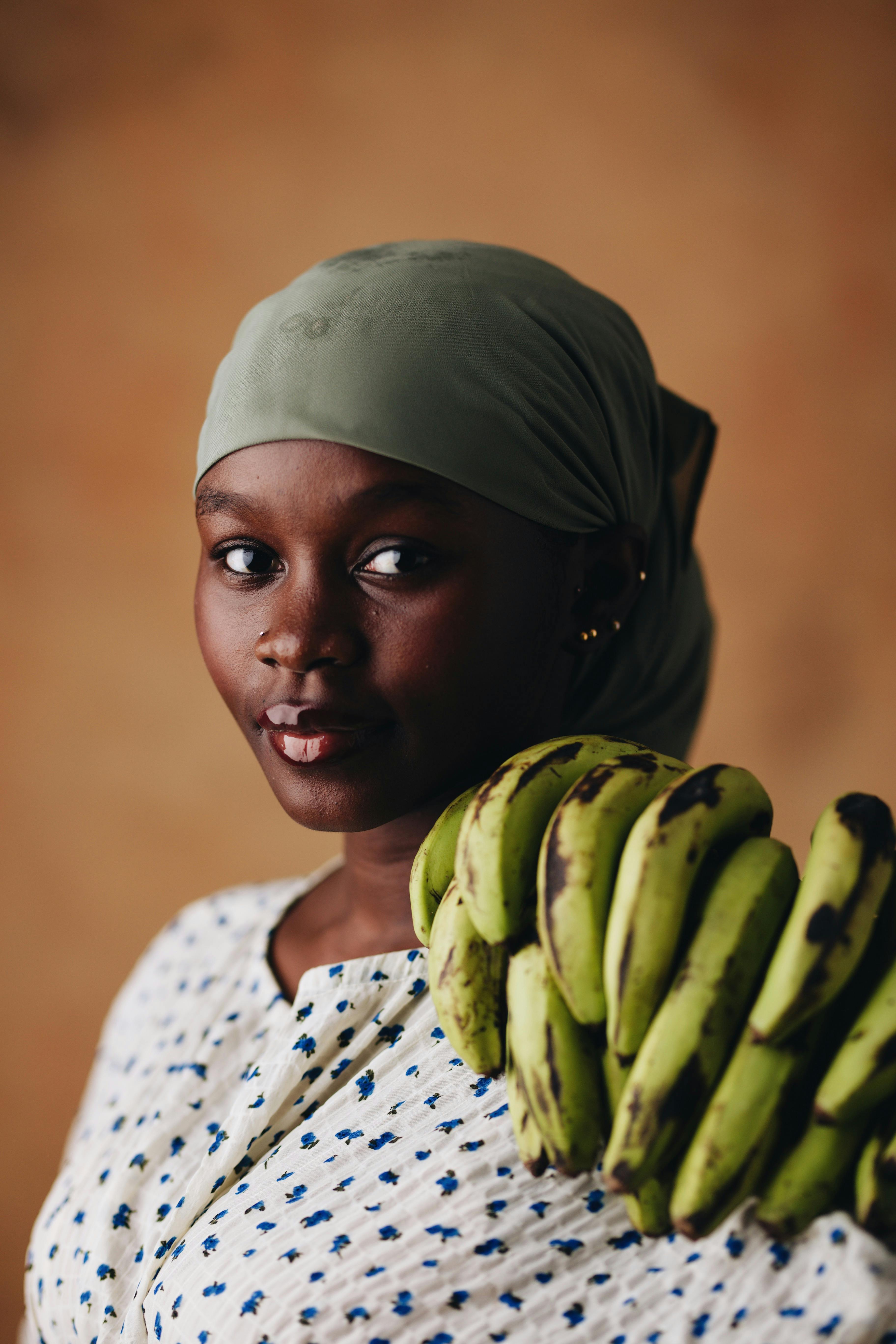 A young woman in a headscarf holds a bunch of green plantains, set against a warm background.