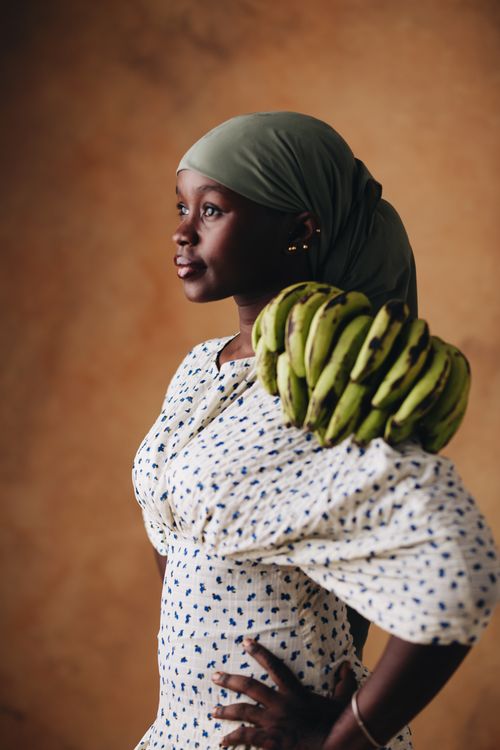 Free A woman in a patterned dress holds a bunch of plantains against a warm backdrop. Stock Photo