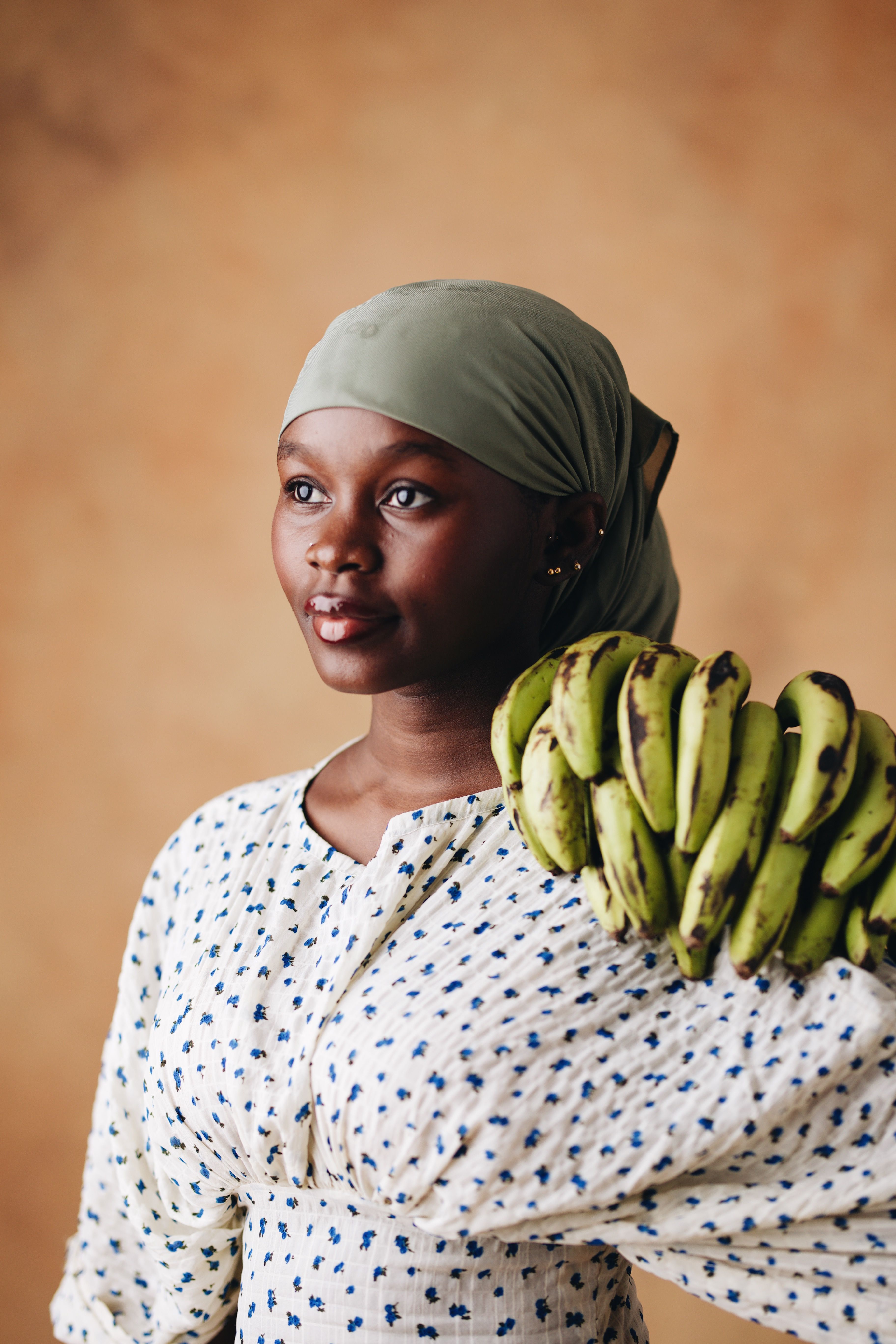 A woman in a headscarf and patterned dress holds green plantains against a warm-toned backdrop.