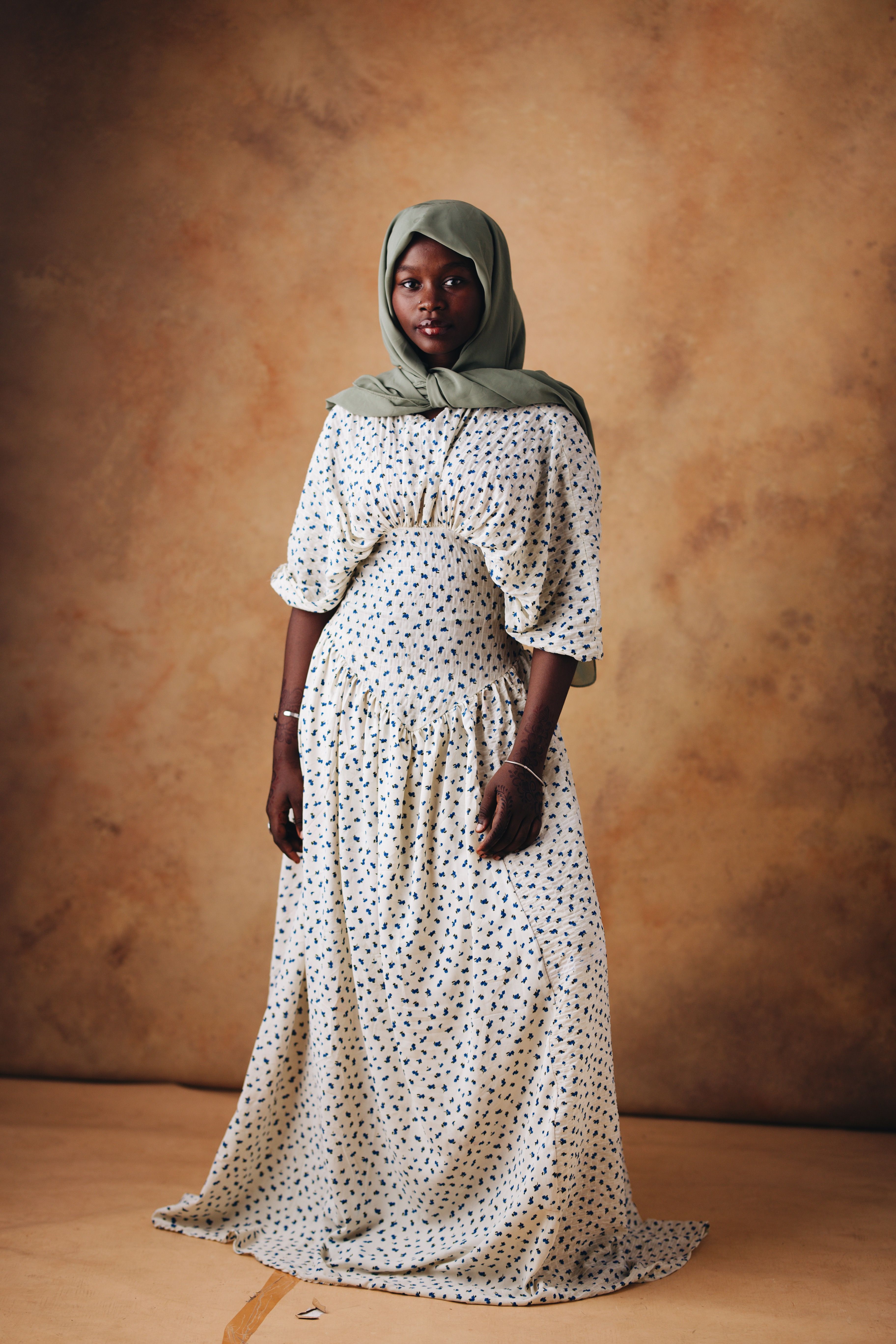 A graceful portrait of a woman posing indoors in a floral dress and headscarf against a neutral backdrop.