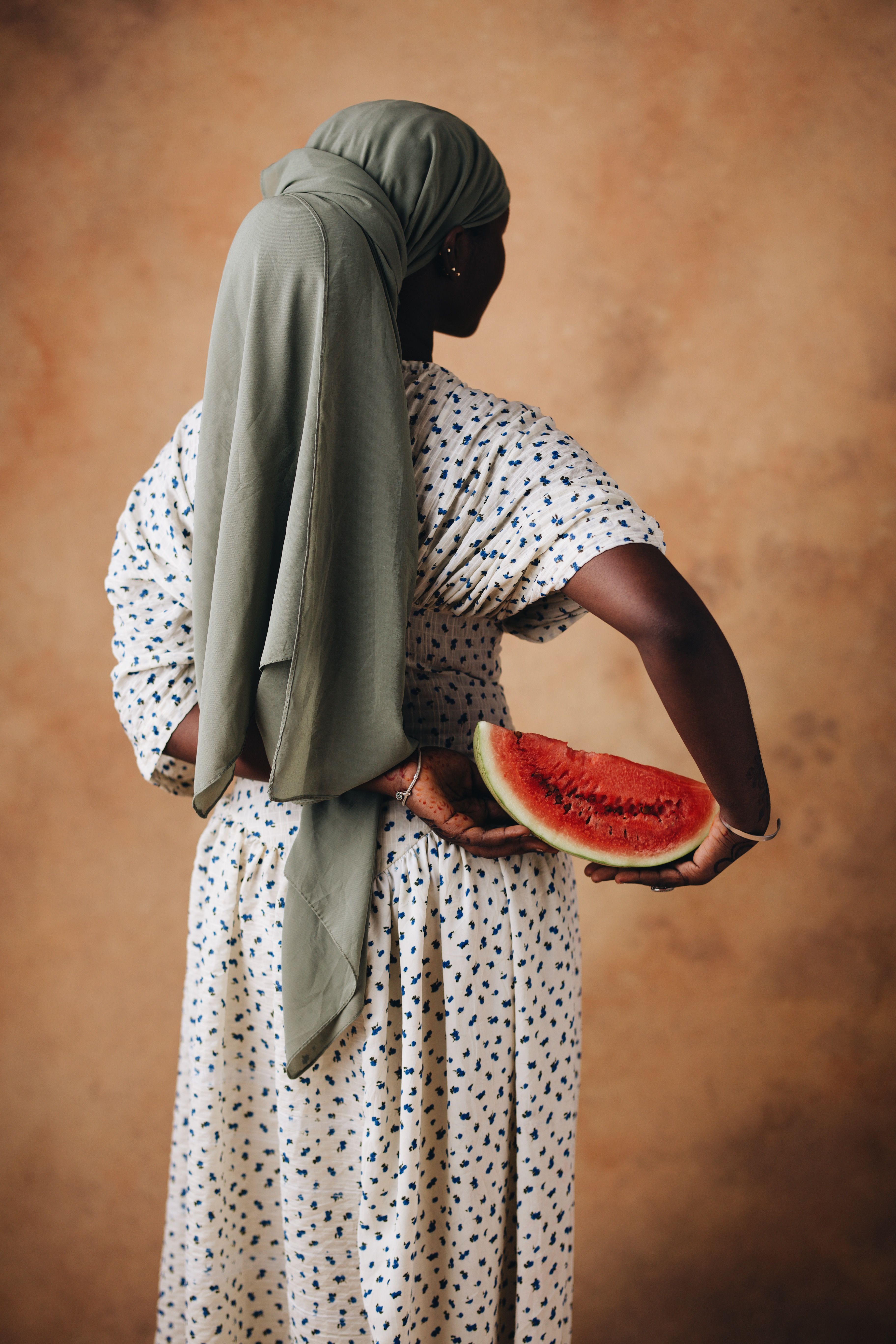 Back view of a woman in a stylish dress holding a slice of watermelon against a warm backdrop.