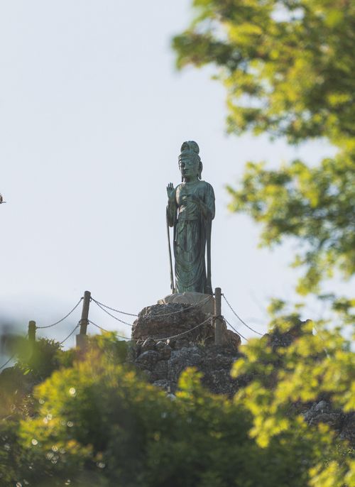 Free A tranquil Buddha statue stands on a rocky hill, surrounded by vibrant green foliage on a sunny day. Stock Photo