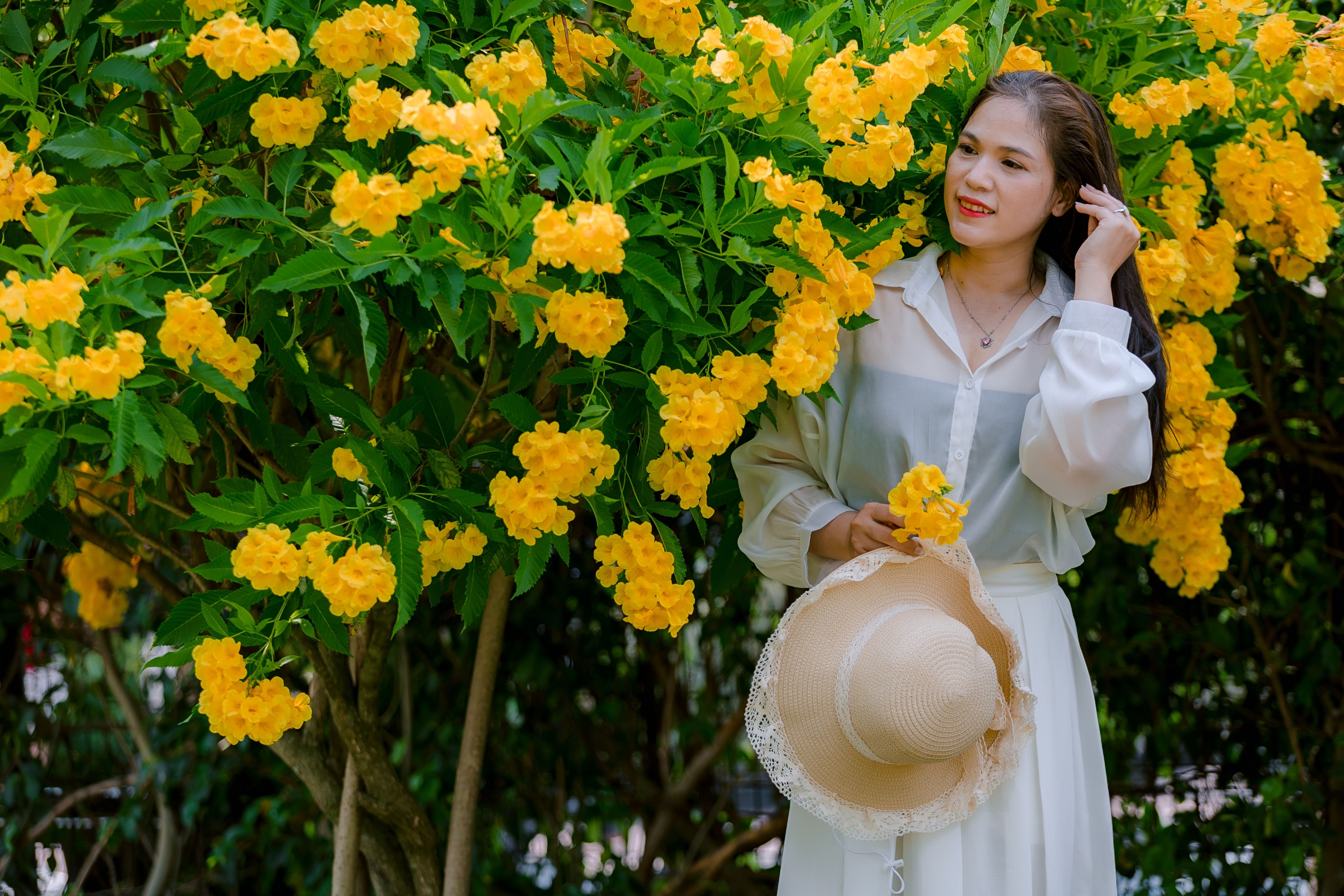 Elegant woman in a white dress stands beside vibrant yellow blossoms holding a straw hat.