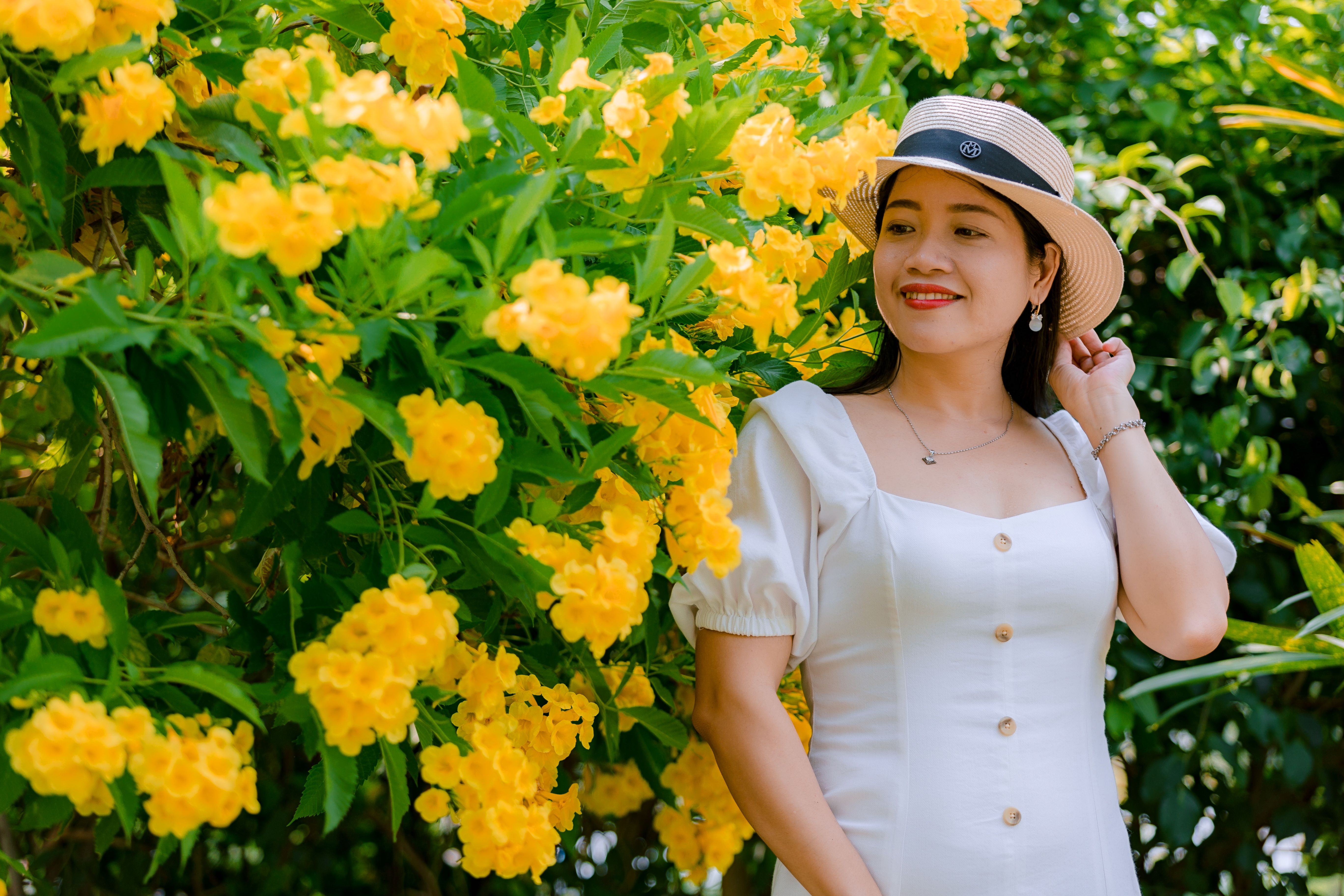 Smiling woman in a white dress and hat poses beside vibrant yellow blossoms outdoors.