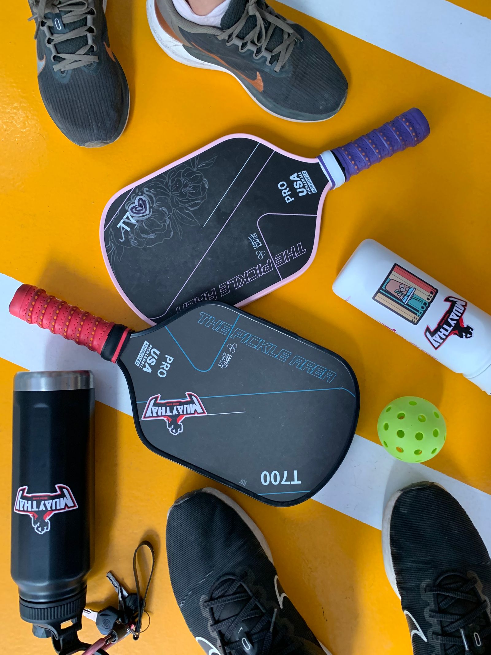 Pickleball paddles, a ball, and court shoes arranged in a flat lay on a court surface
