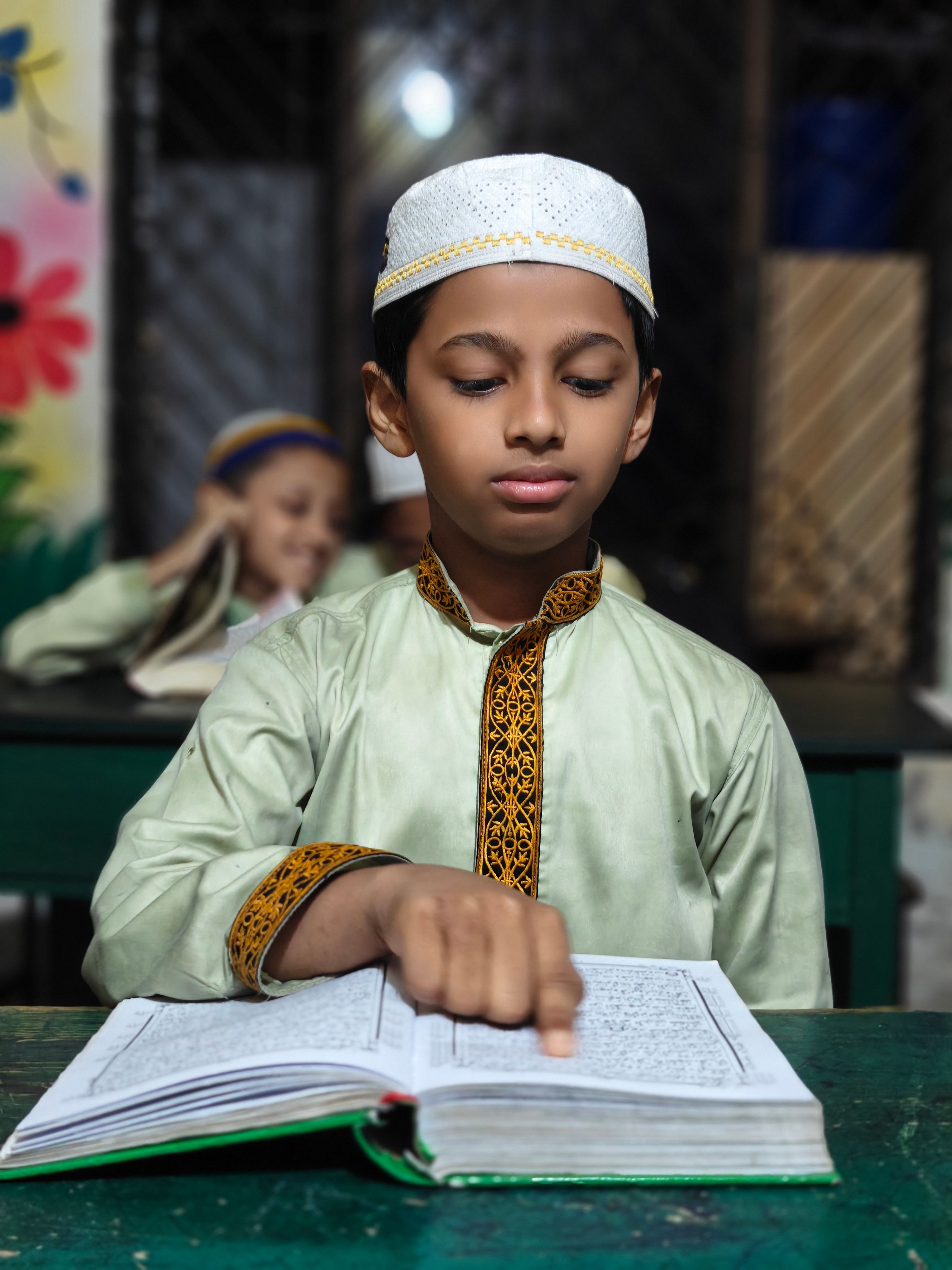 A young boy in traditional attire reads a religious book in a classroom setting.