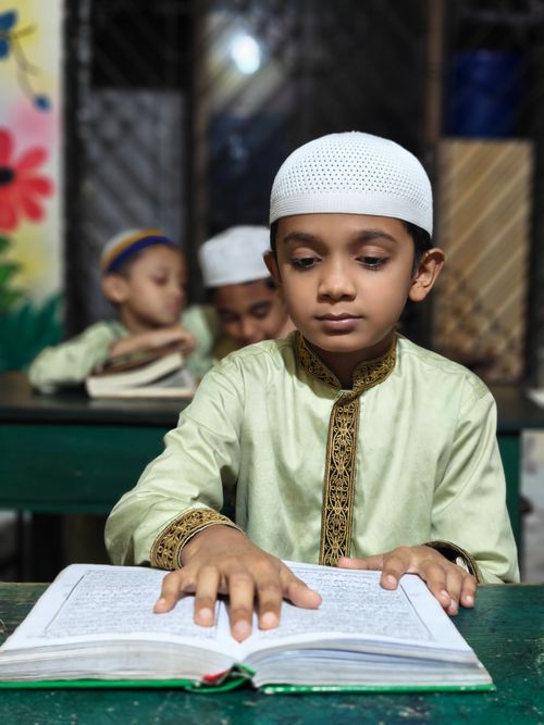 Free A young boy in a traditional outfit reading the Quran in a classroom setting. Stock Photo