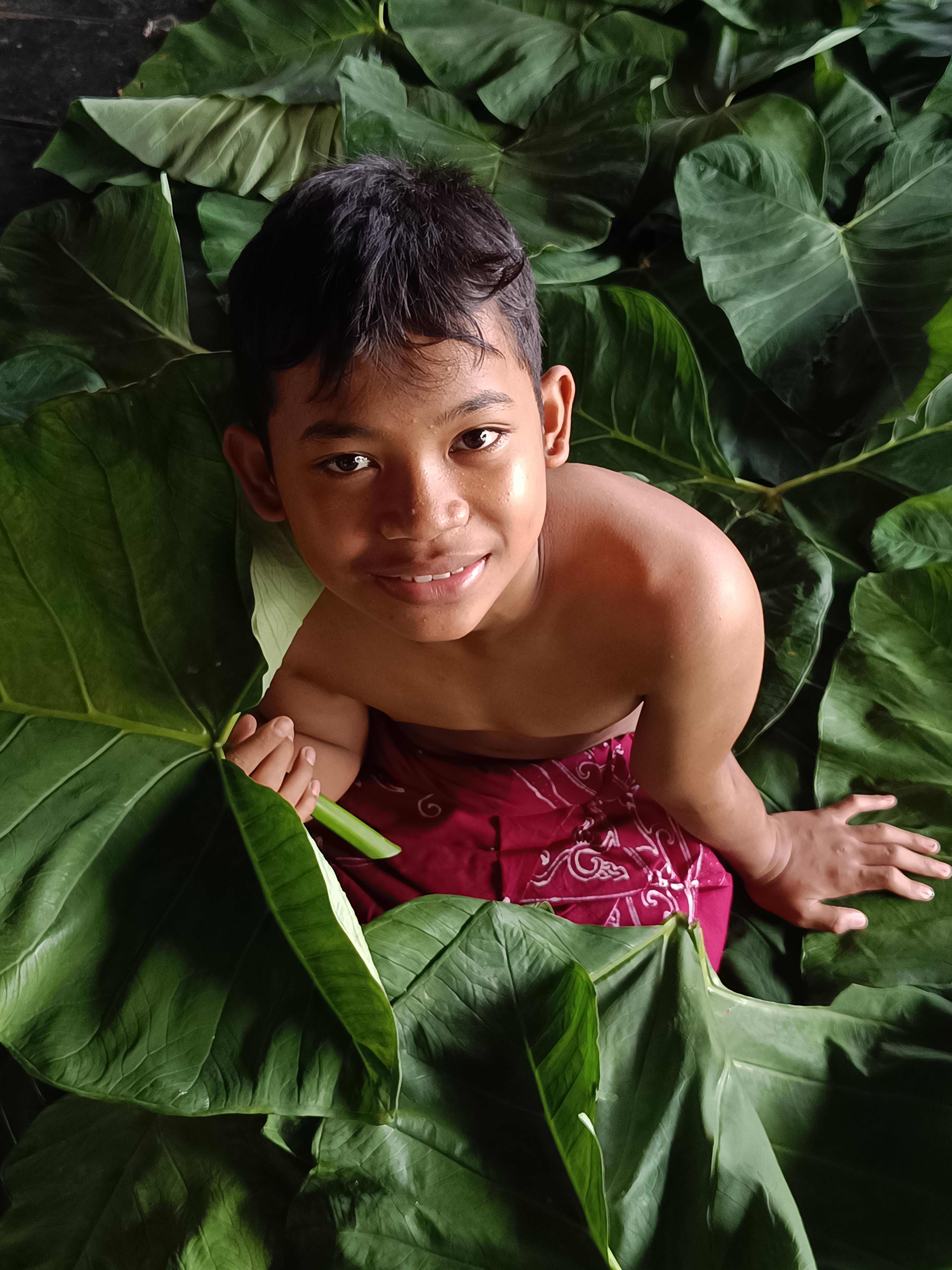 A cheerful young boy smiles while surrounded by large green leaves, symbolizing nature's embrace.