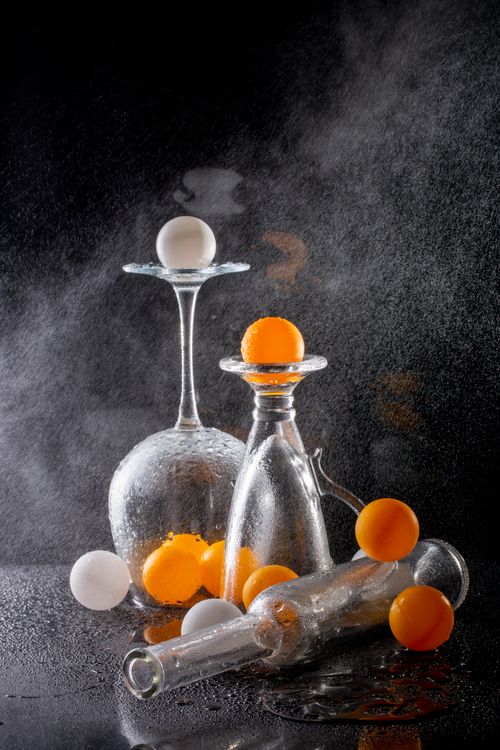 Free Creative still life arrangement featuring orange and white ping pong balls with glassware against a dramatic backdrop. Stock Photo
