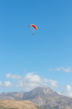 Vibrant paraglider flies high above a scenic mountain landscape with clear blue skies.