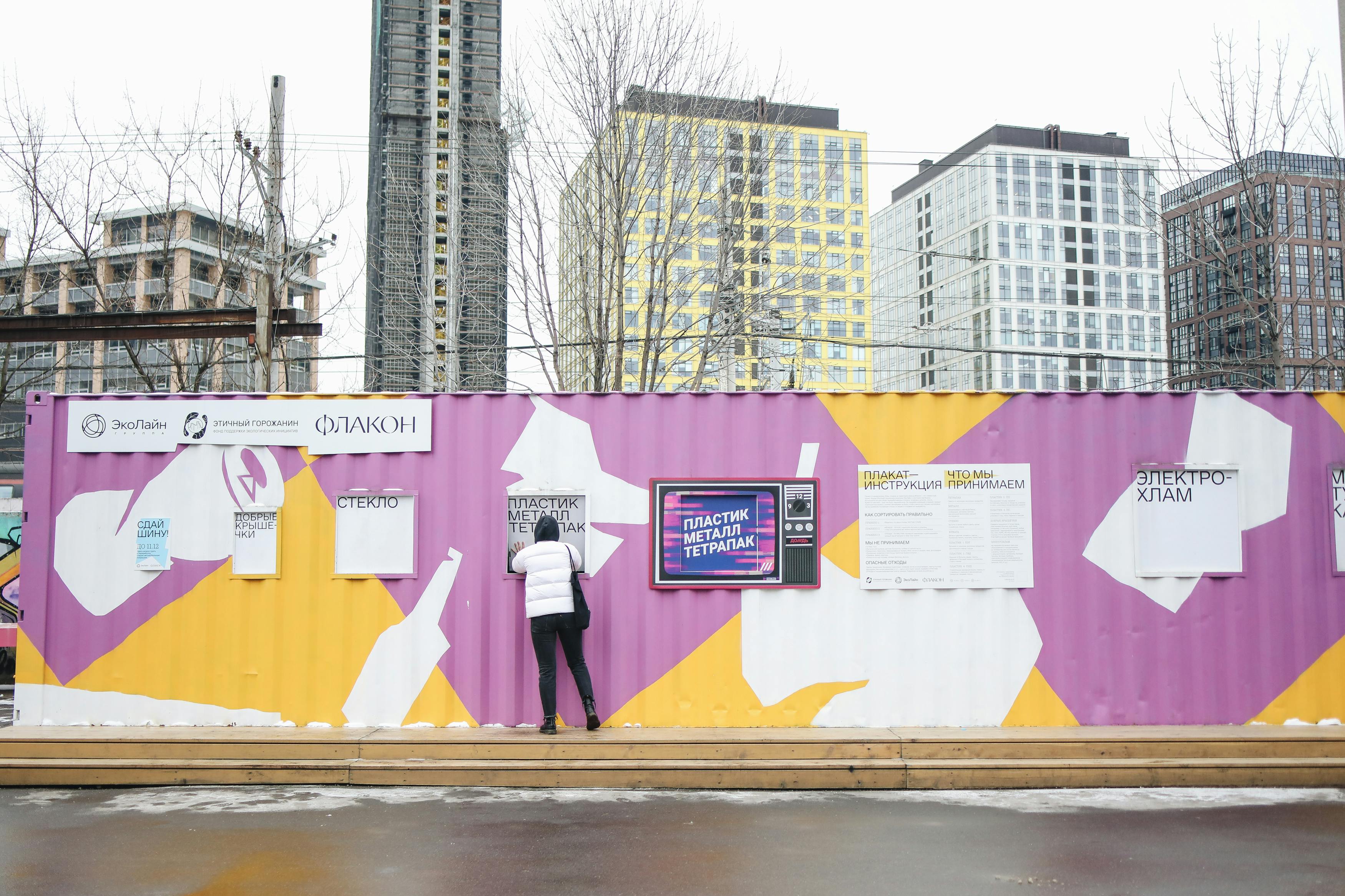 在五颜六色的容器和城市建筑背景下，一个人将废物存放在一个充满活力的城市回收站