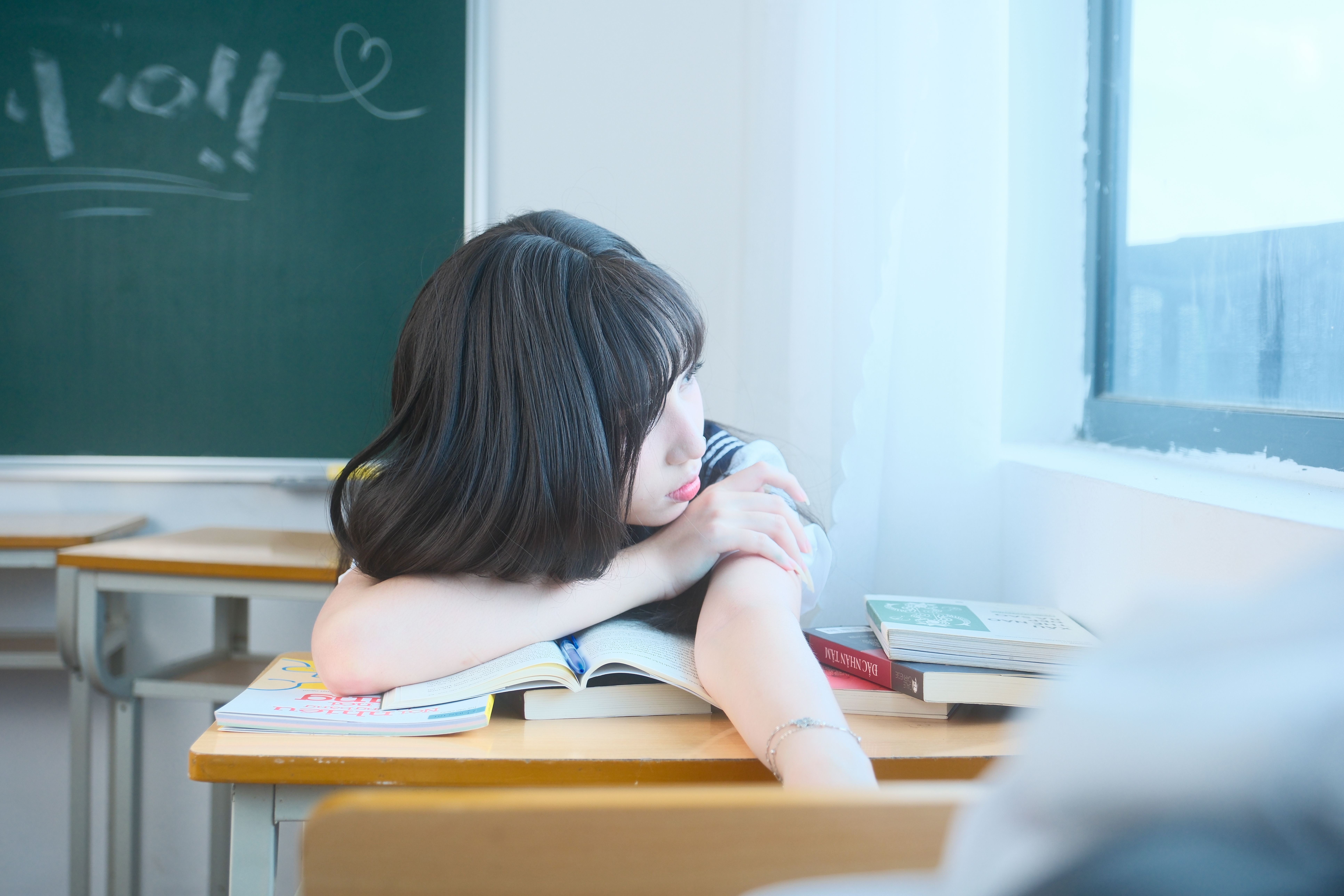 A student daydreaming beside the window in a Vietnamese classroom, surrounded by books.