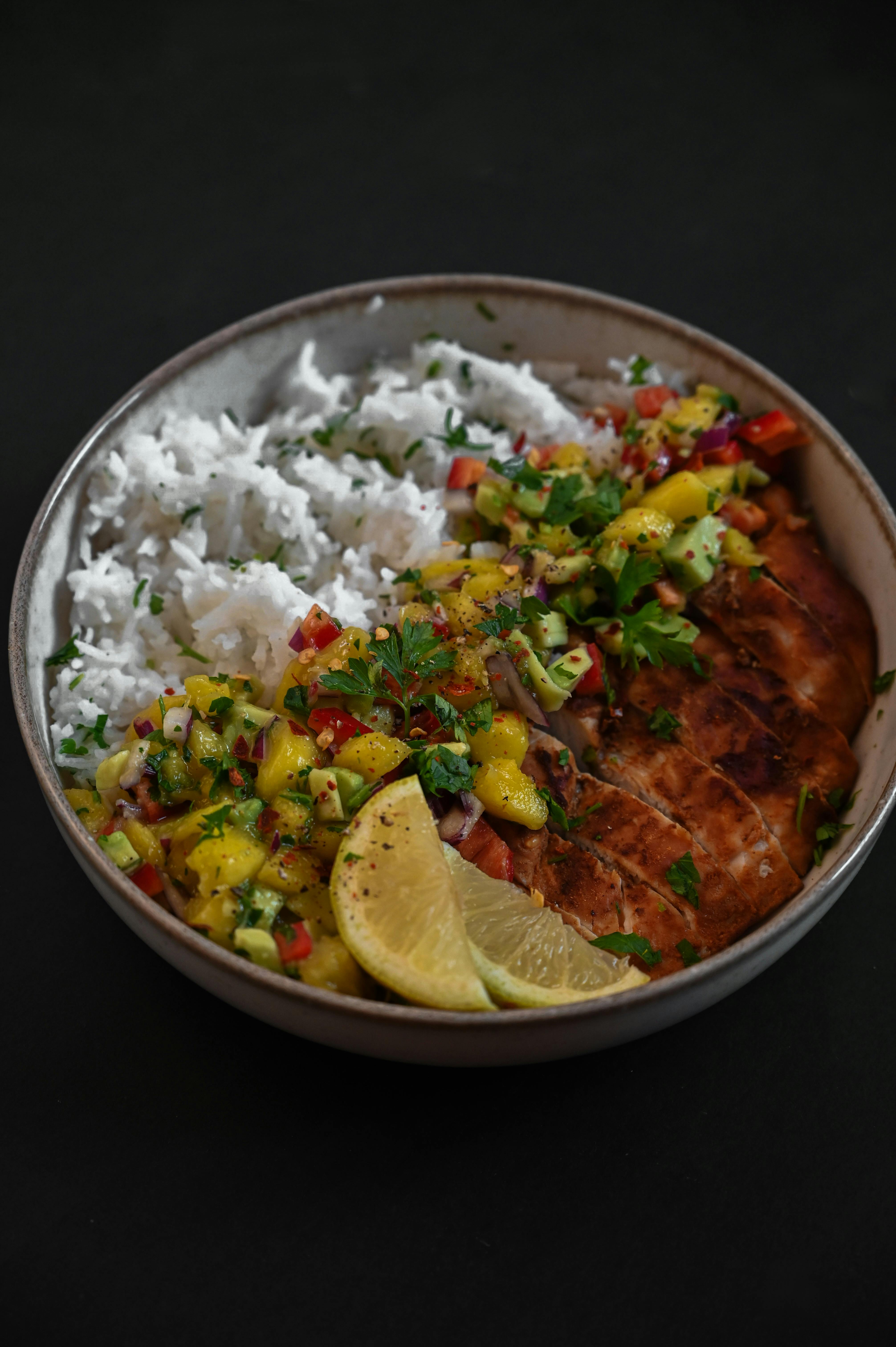 A bowl of coconut rice topped with golden-brown chicken thighs in creamy coconut sauce, colorful mango slaw, and fresh cilantro garnish