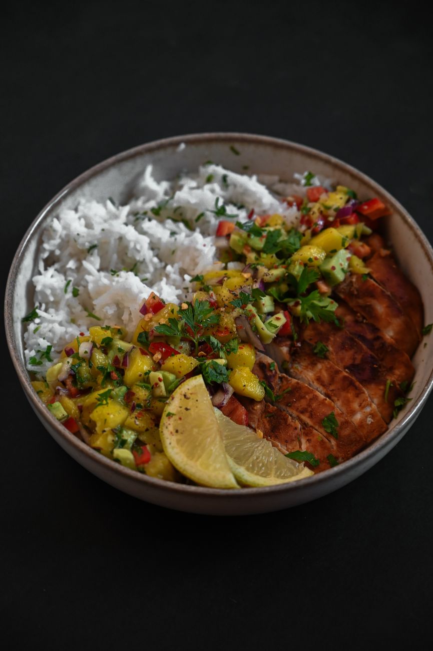 A bowl of coconut rice topped with golden-brown chicken thighs in creamy coconut sauce, colorful mango slaw, and fresh cilantro garnish