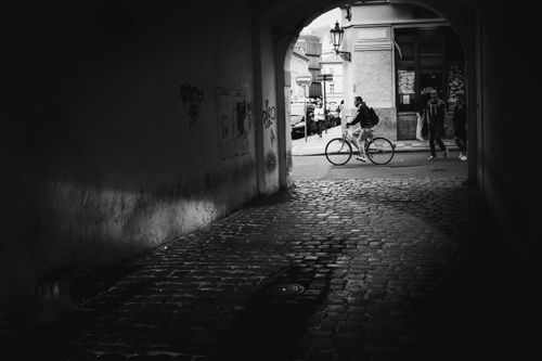 Free A black and white photo capturing a man cycling through a historic archway in Prague's old city. Stock Photo