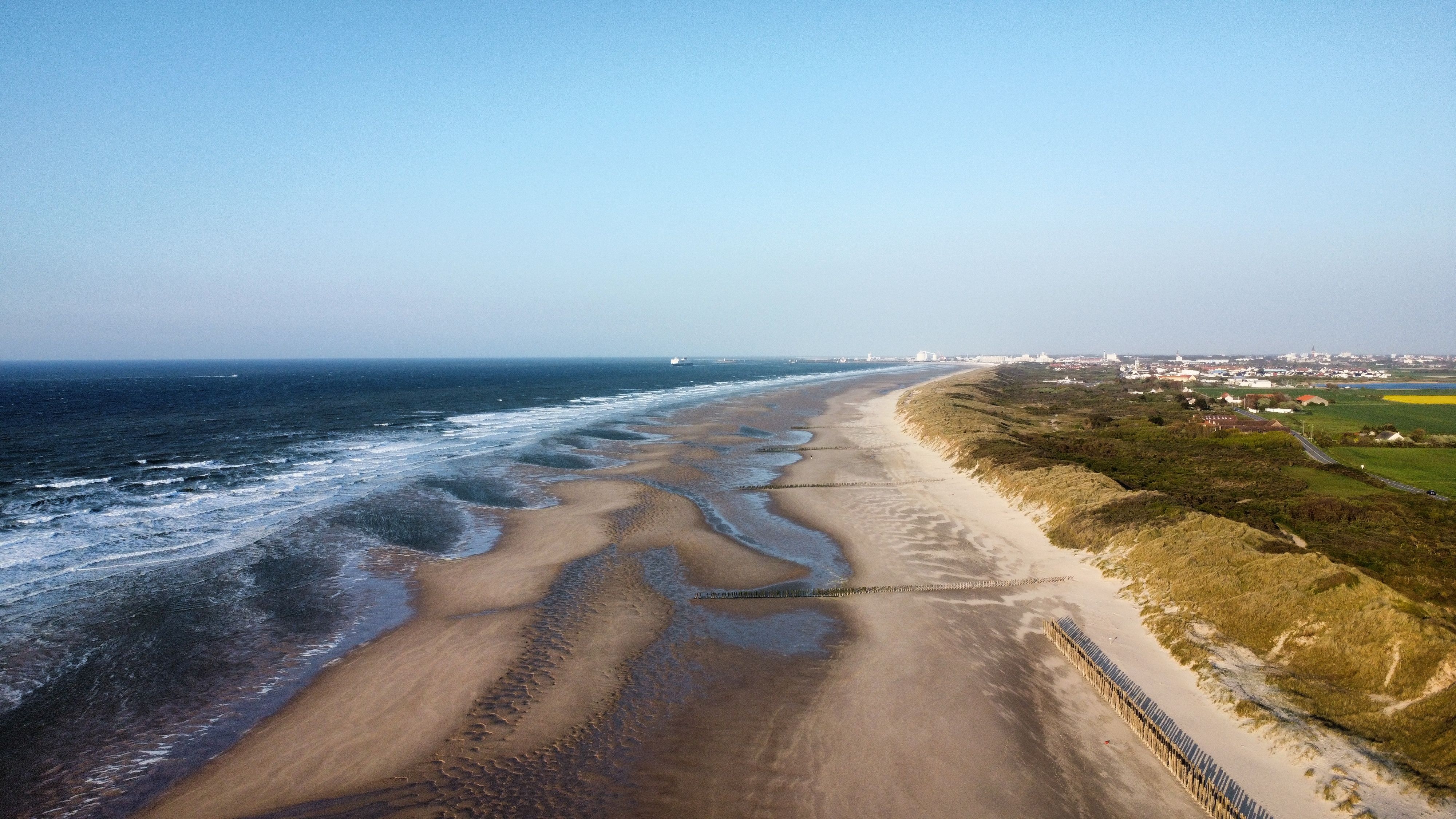 Aerial view of a long sandy coastline with rolling waves, dunes and a distant coastal town